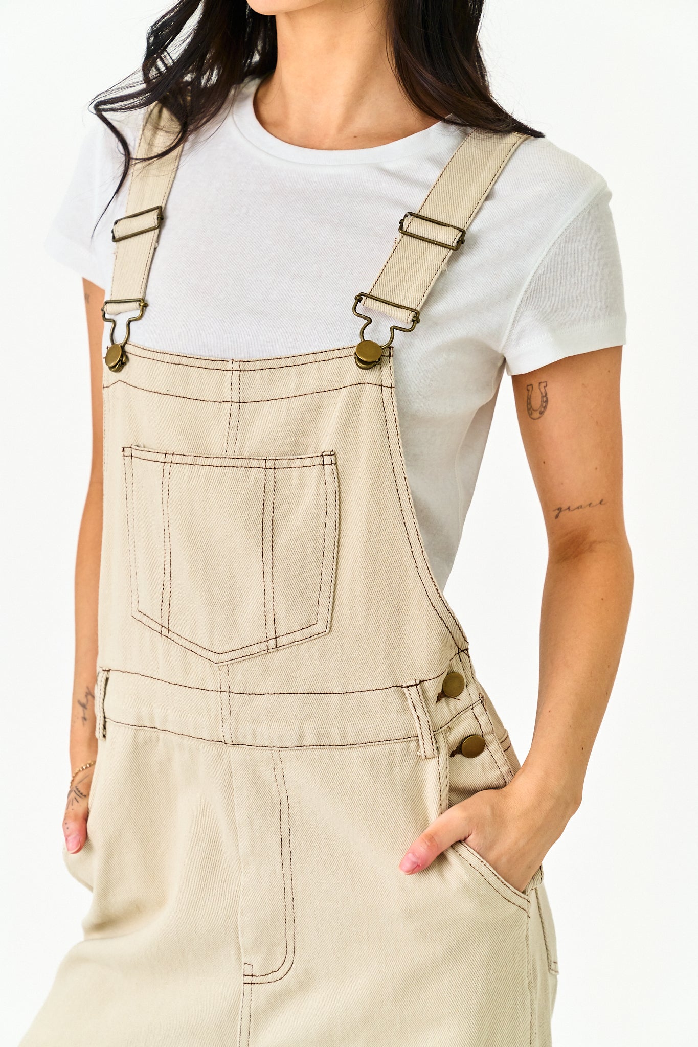 A person wearing light beige denim-style overalls over a white short-sleeve T-shirt, shown from the neck to the hips against a plain light background.