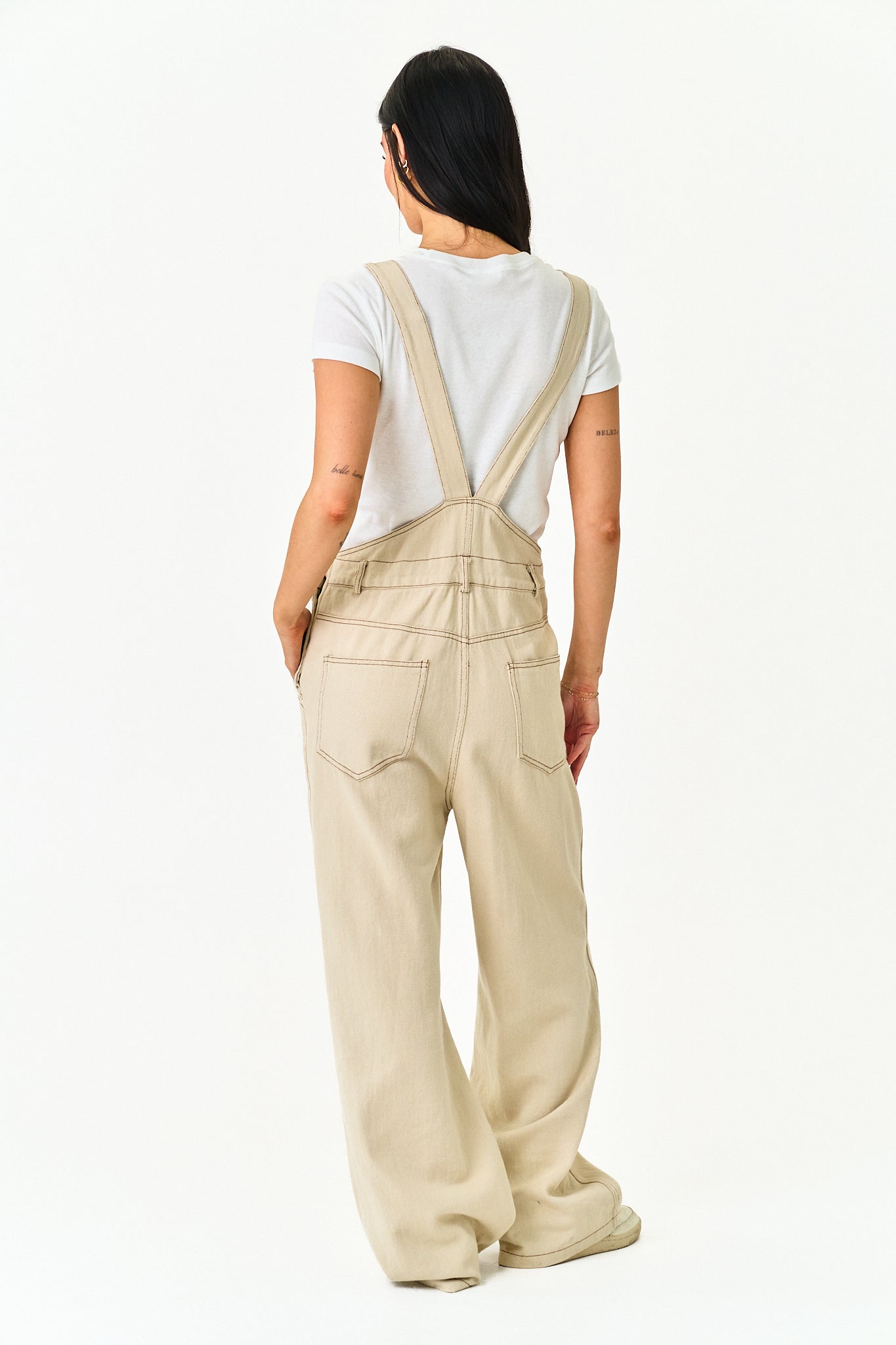 A woman viewed from behind wearing beige wide-leg overalls with a white t-shirt, standing against a plain white studio background.