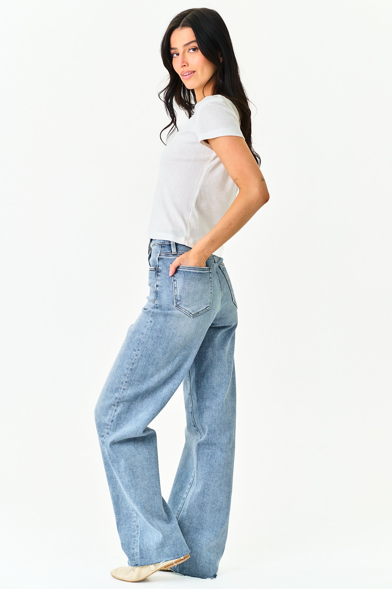 A woman modeling a pair of light-wash, wide-leg denim jeans with a white t-shirt, standing against a plain white studio background.