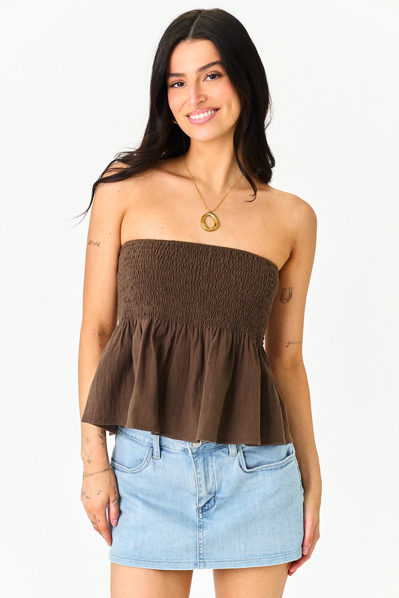 A woman modeling a brown strapless smocked peplum top paired with a light blue denim mini skirt, set against a plain white studio background.