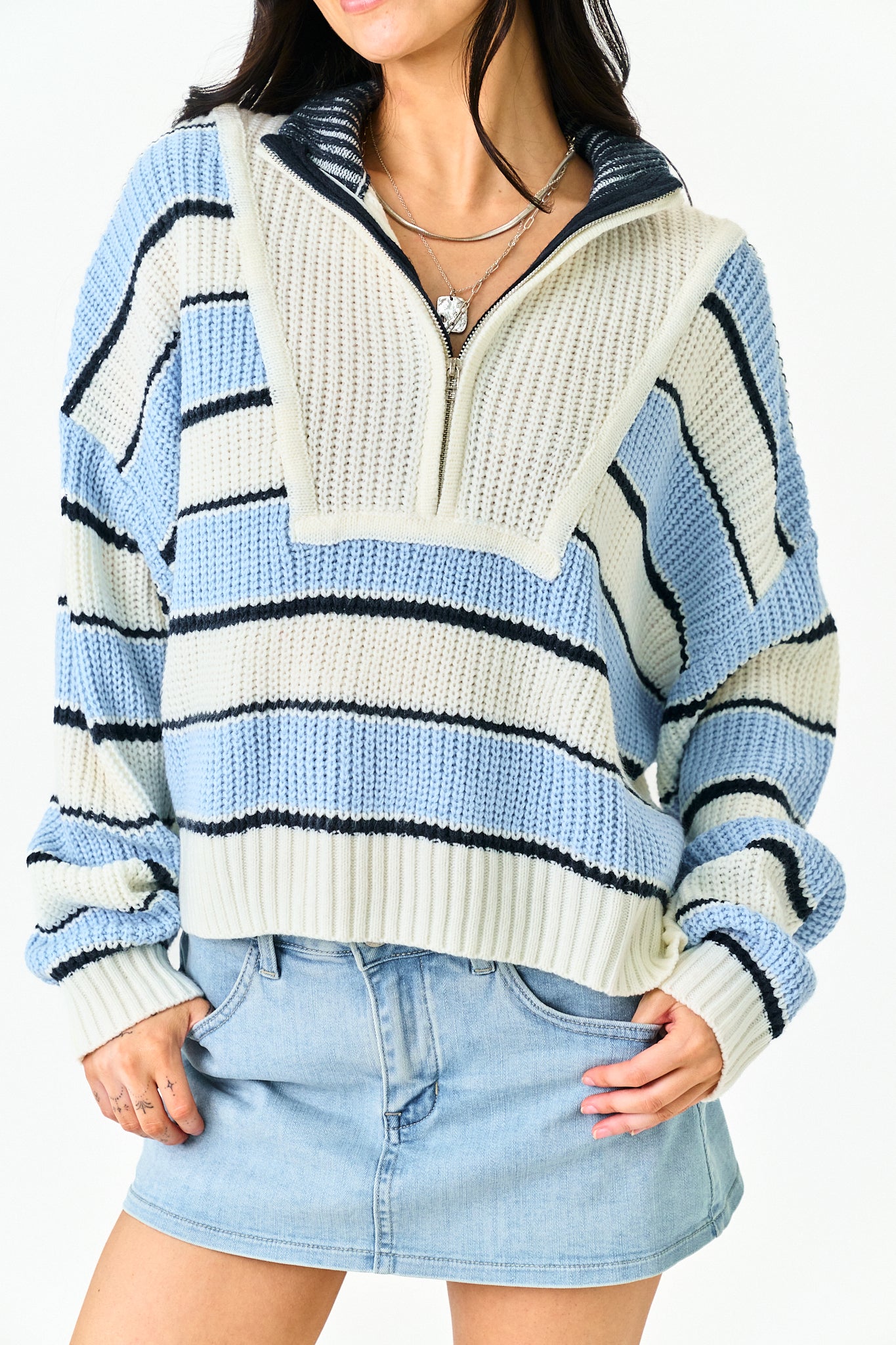A woman wearing a chunky knit quarter-zip sweater with horizontal stripes in light blue, cream, and black, paired with a light blue denim mini skirt.