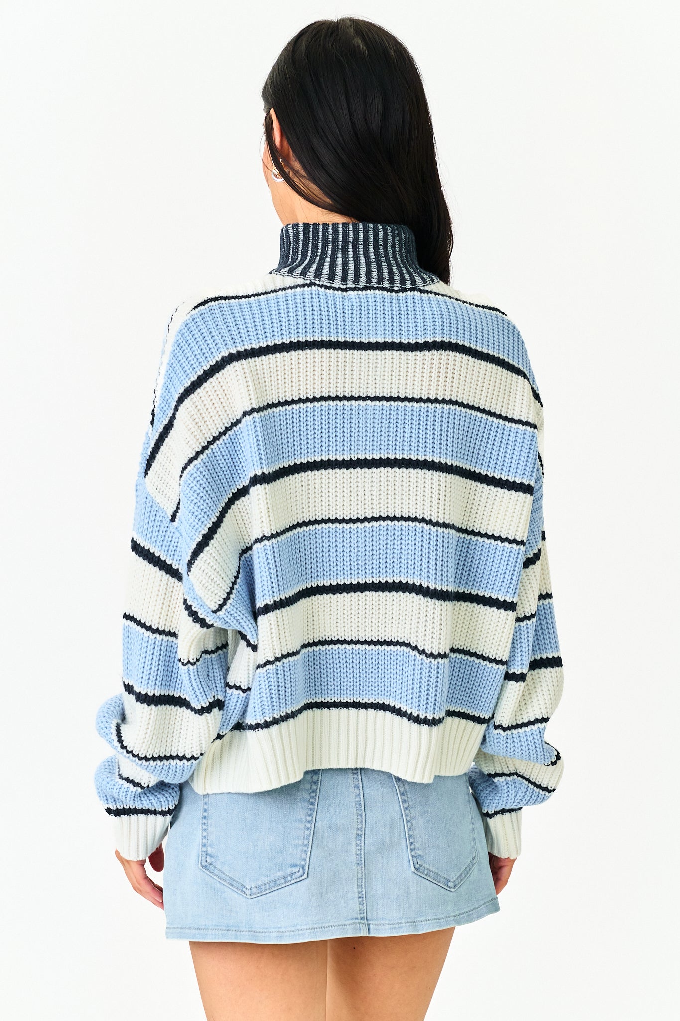 A woman shown from the back wearing a light blue, white, and black striped knit turtleneck sweater and a light blue denim mini skirt, against a plain white studio background.
