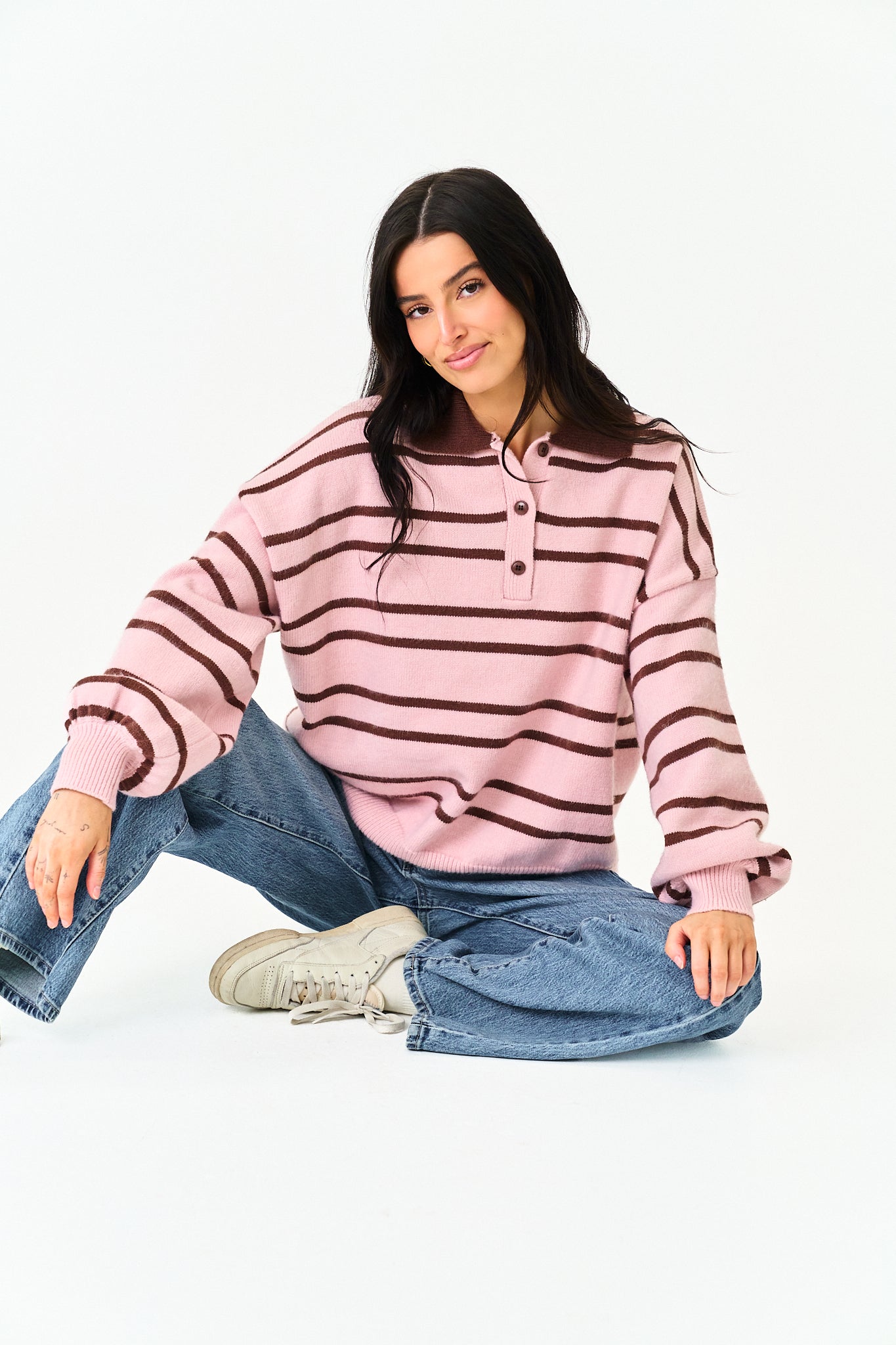 A woman sitting cross-legged on a white studio floor wearing a pink and dark brown striped long-sleeve henley sweater, blue jeans, and white sneakers.