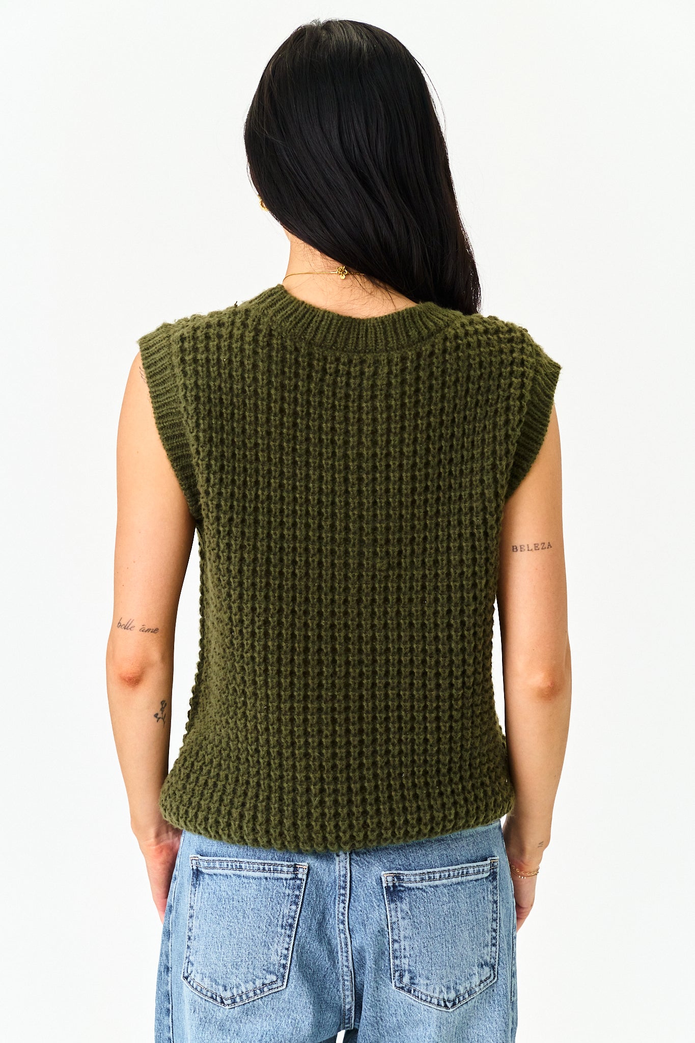 A woman viewed from the back wearing a sleeveless olive green ribbed knit top and light blue jeans against a plain white background.