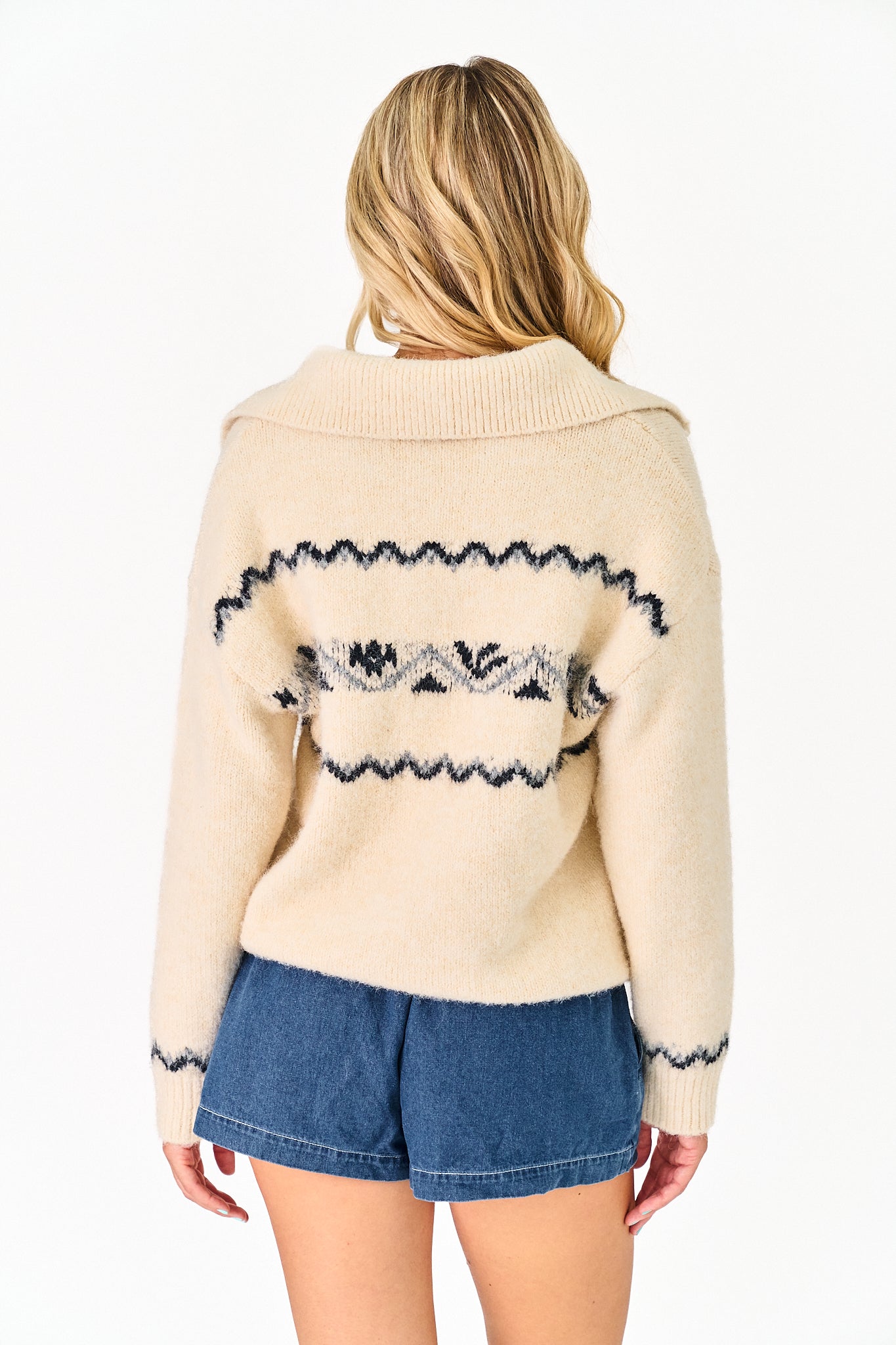 A woman viewed from the back wearing a cream knit sweater with a dark navy and gray patterned horizontal stripe across the upper back, paired with blue denim shorts, against a plain white background.