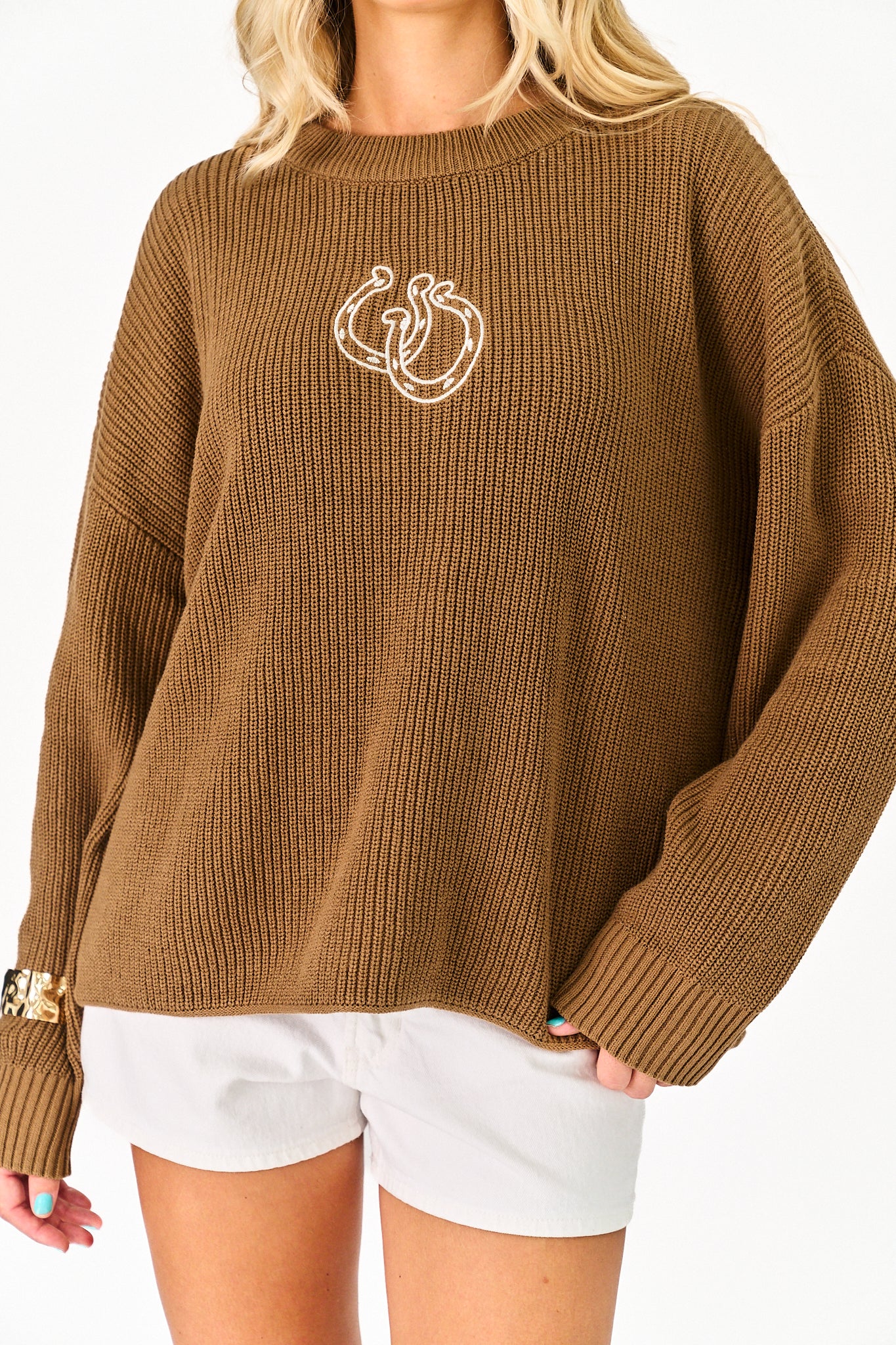 A woman wearing an oversized ribbed knit sweater in a warm camel brown color, featuring a white line-art graphic of two snakes on the chest, paired with white shorts.
