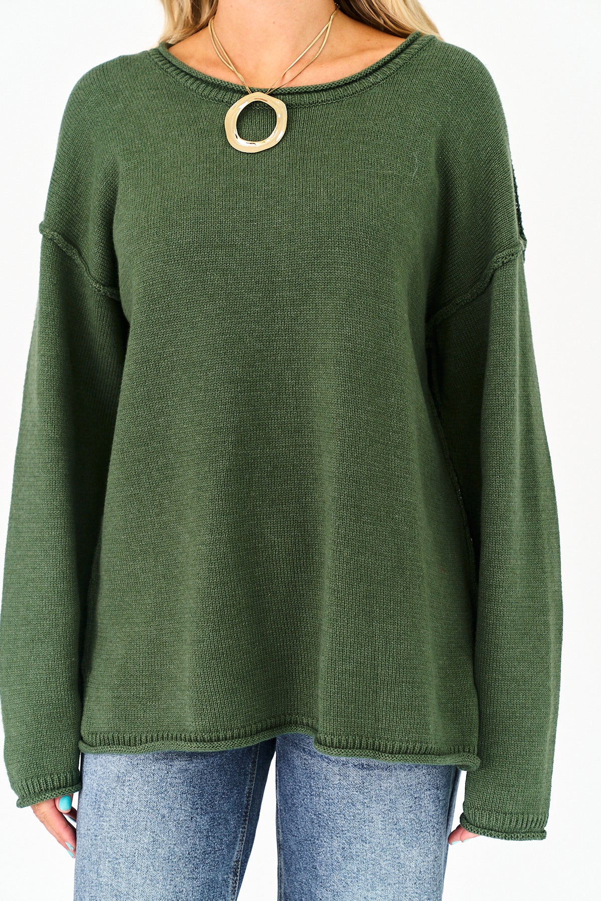 An olive green knit sweater with a wide crew neckline and a large gold circular pendant necklace worn by a model, paired with blue jeans.