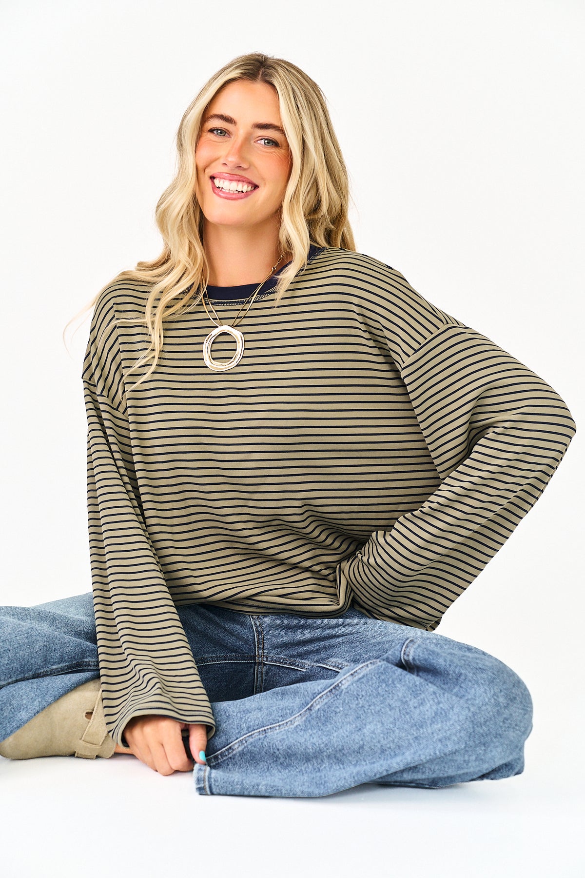 A smiling blonde woman sits cross-legged on a white studio floor wearing a loose, long-sleeve striped top in olive green and beige, blue jeans, and a large circular pendant necklace.