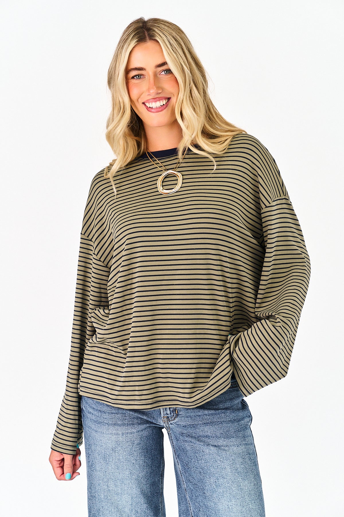 A smiling blonde woman wearing a loose, long-sleeve striped knit top in olive green and black, paired with blue jeans, against a plain white studio background.