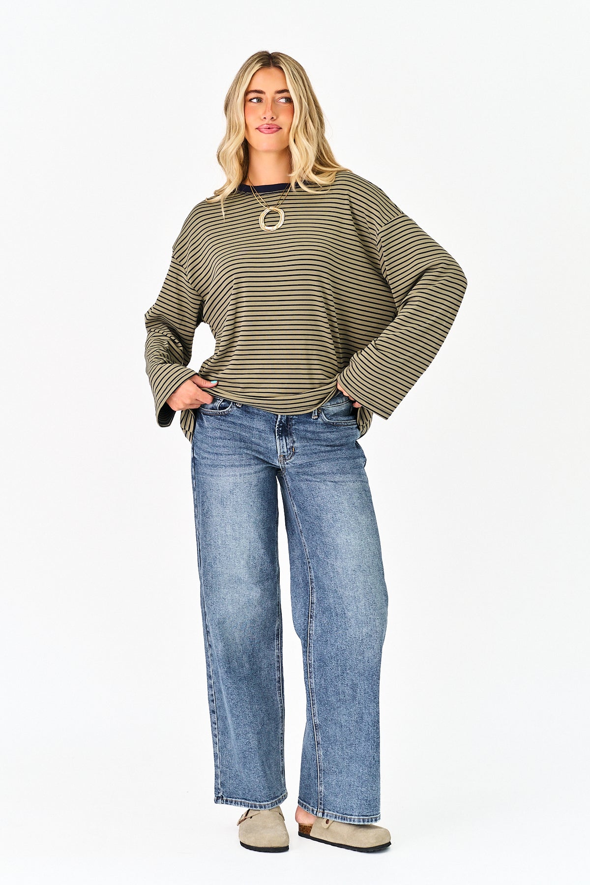 A woman modeling a loose-fitting, olive green and beige horizontally striped long-sleeve top with wide bell sleeves, paired with blue jeans and beige shoes, against a plain white background.