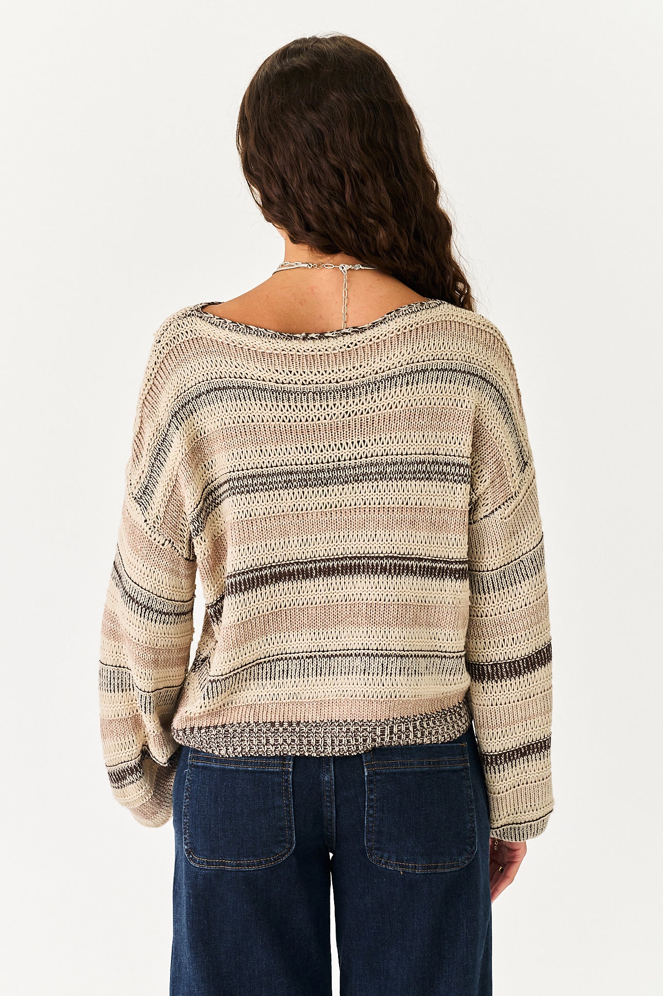 A woman viewed from behind wearing a loose, long-sleeve knit sweater with horizontal beige, brown, and black stripes, paired with dark blue jeans against a plain white background.