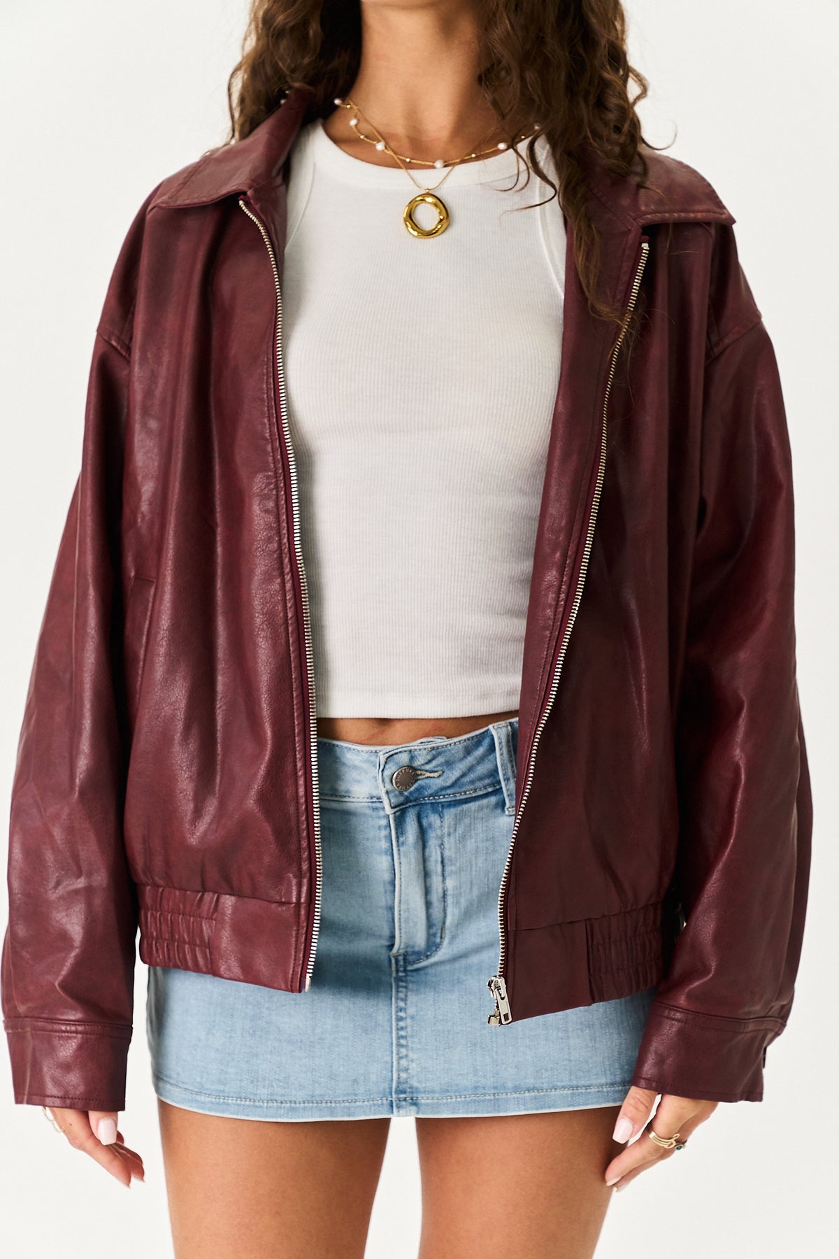 A woman wearing a burgundy faux-leather zip-up jacket over a white crop top and a light blue denim mini skirt, shown from the neck down against a plain white background.