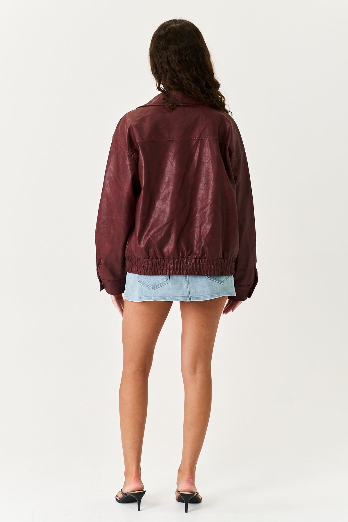 A woman shown from the back wearing a dark burgundy faux-leather jacket, a light blue denim mini skirt, and black mule heels against a plain white studio background.
