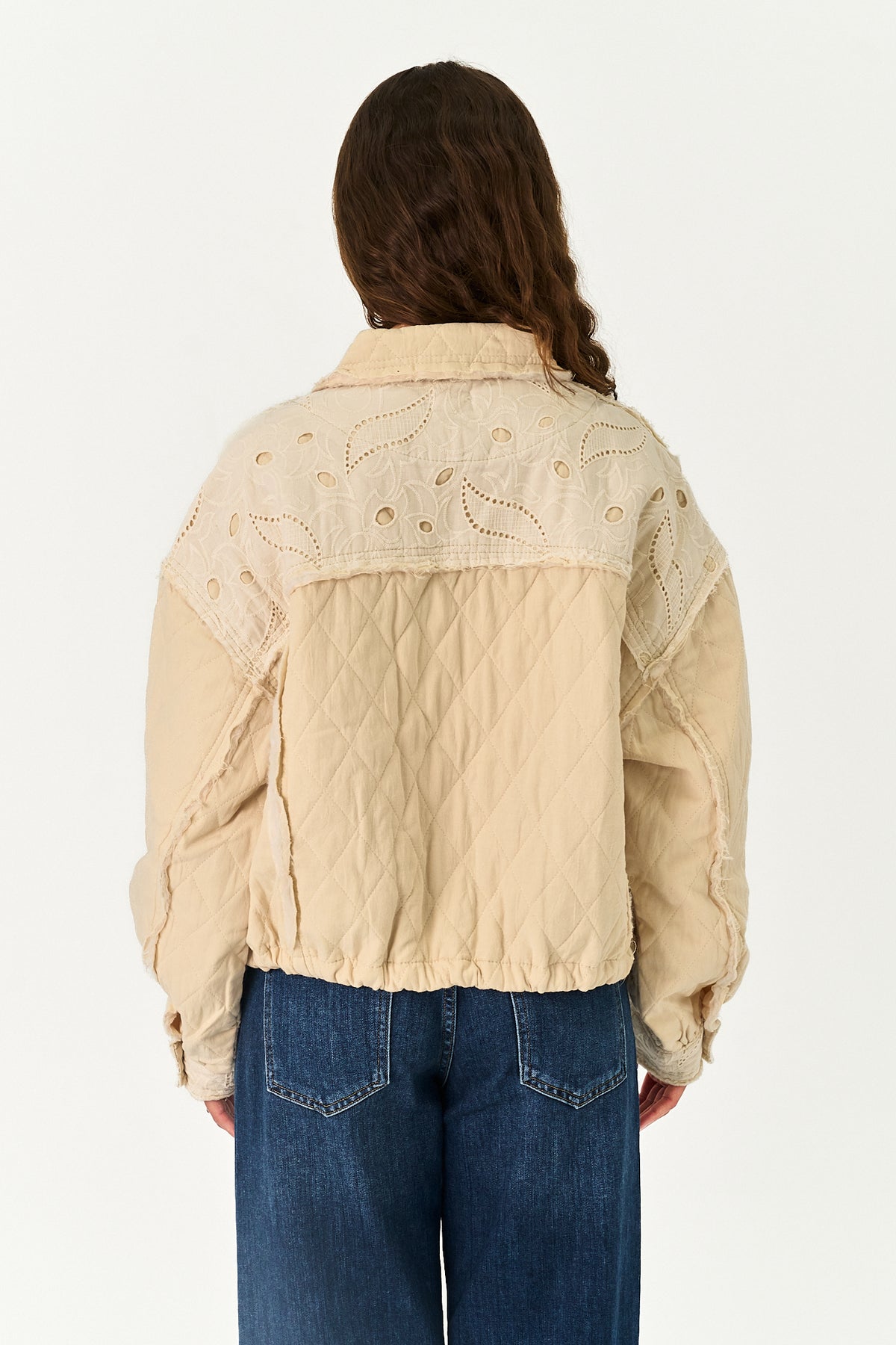 A model viewed from the back wearing a beige quilted jacket with embroidered details on the shoulders and sleeves, paired with blue jeans, against a plain white background.