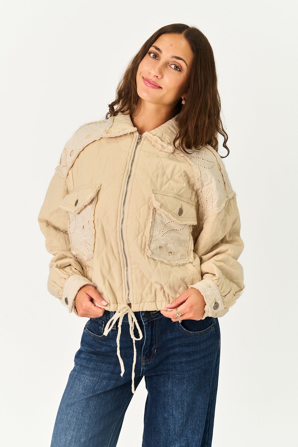 A woman wearing a beige quilted jacket with lace applique details and blue jeans, standing against a plain white studio background.