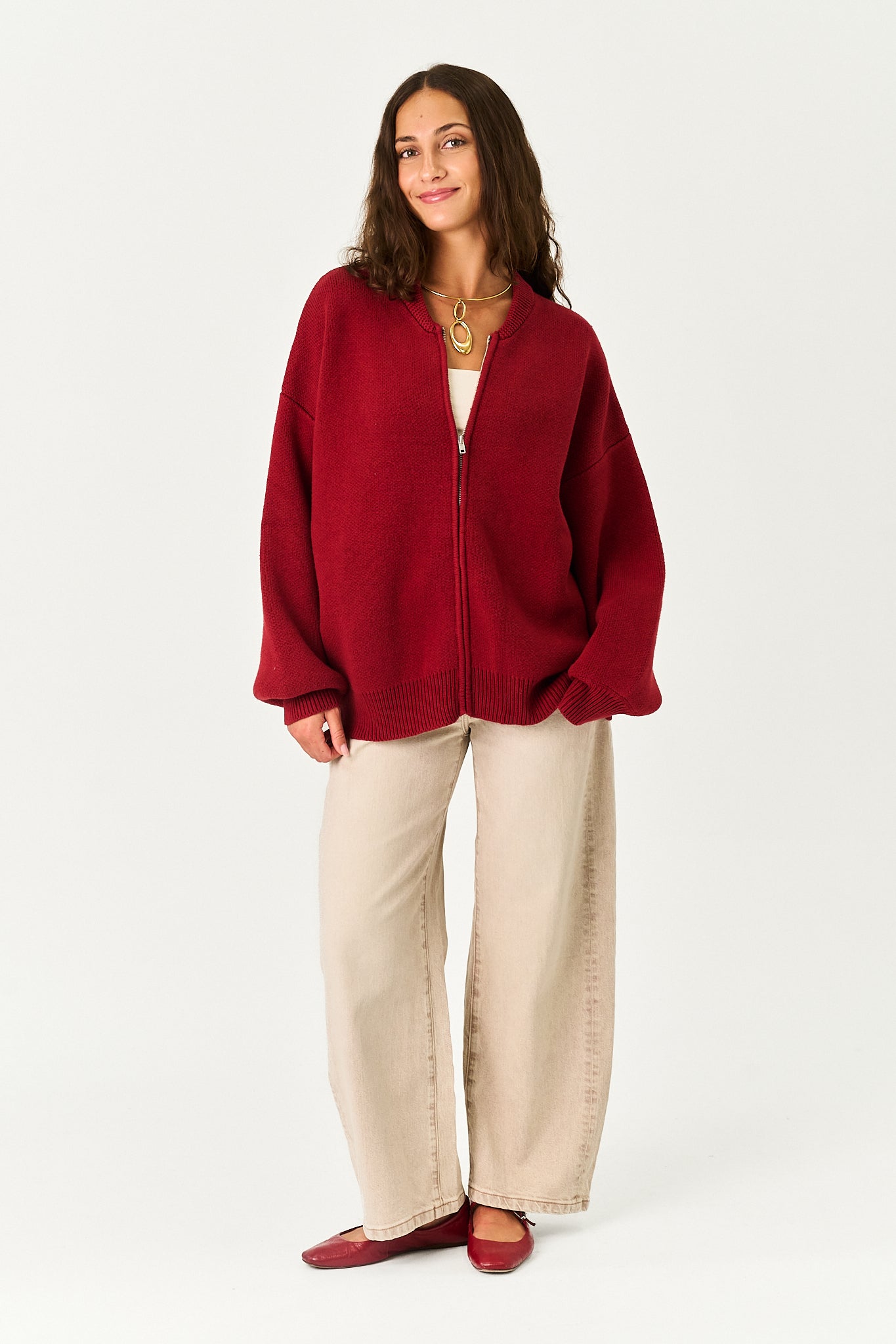A woman modeling an oversized deep red zip-up cardigan with wide beige pants and red flats, standing against a plain light gray studio background.