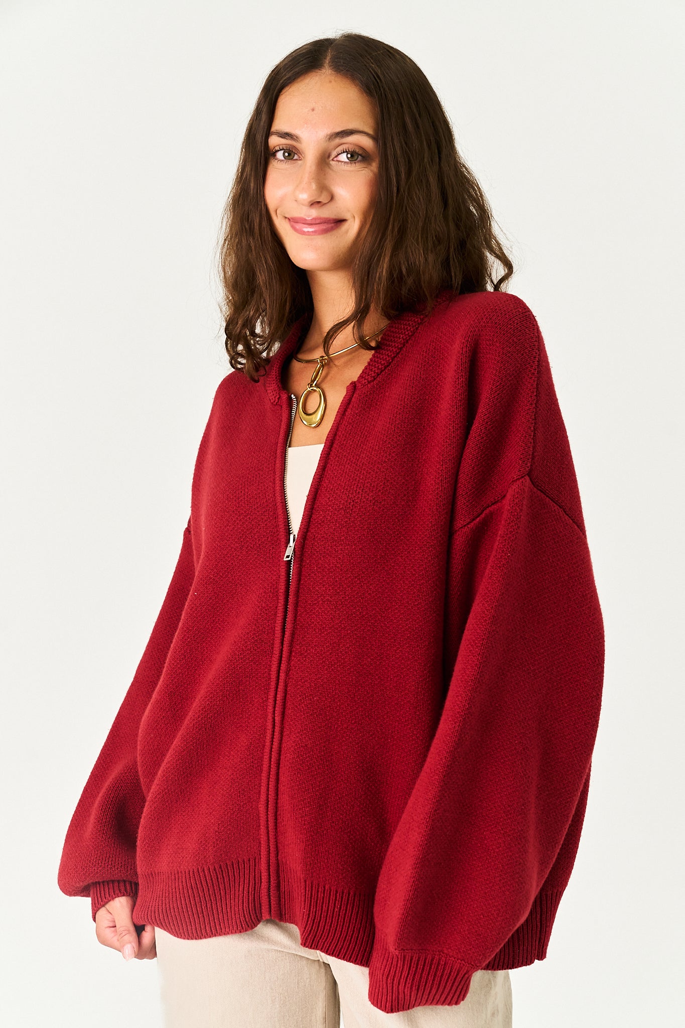 A woman wearing an oversized, deep red zip-up knit cardigan with voluminous balloon sleeves, styled over a white top and beige pants, set against a plain light gray background.