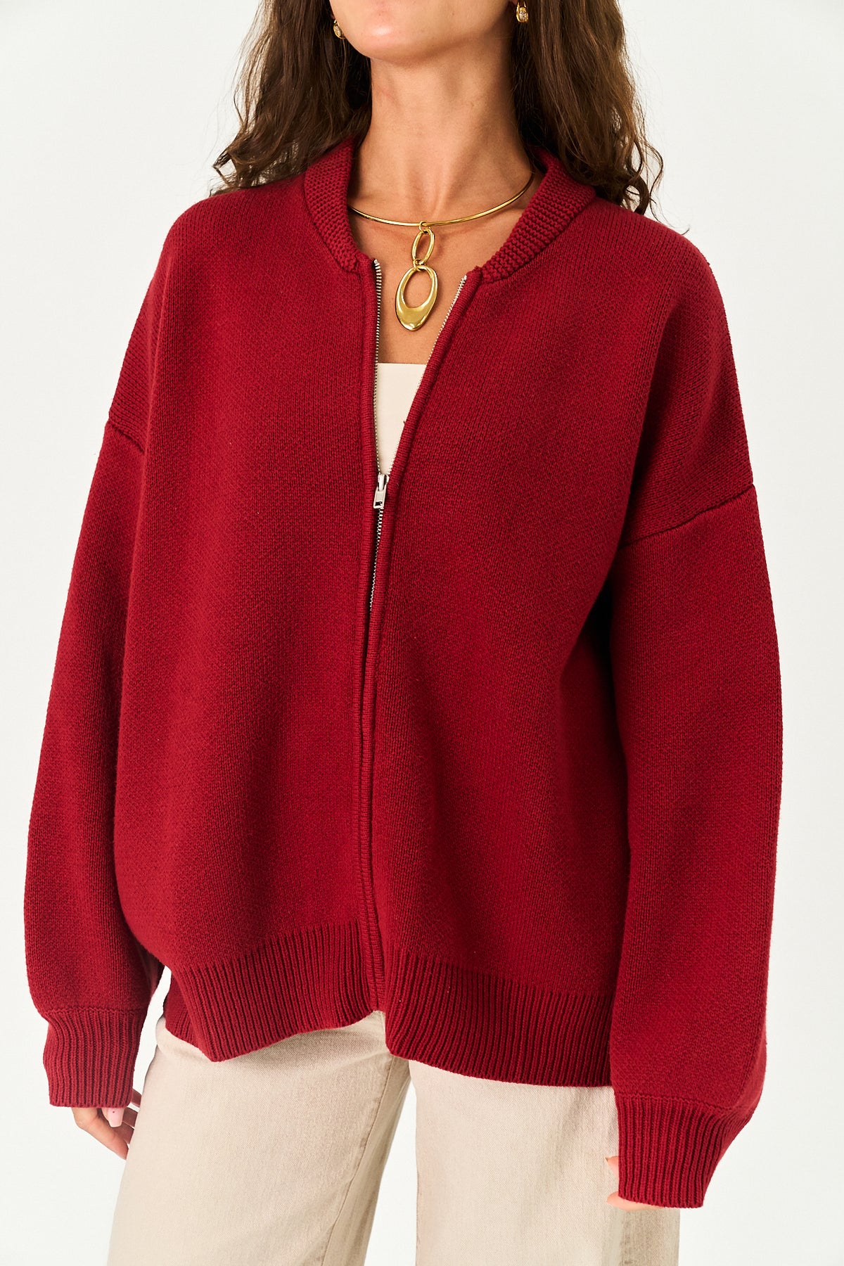 A woman wearing an oversized red zip-up knit cardigan sweater with ribbed collar, cuffs, and hem, layered over a white top and cream pants.
