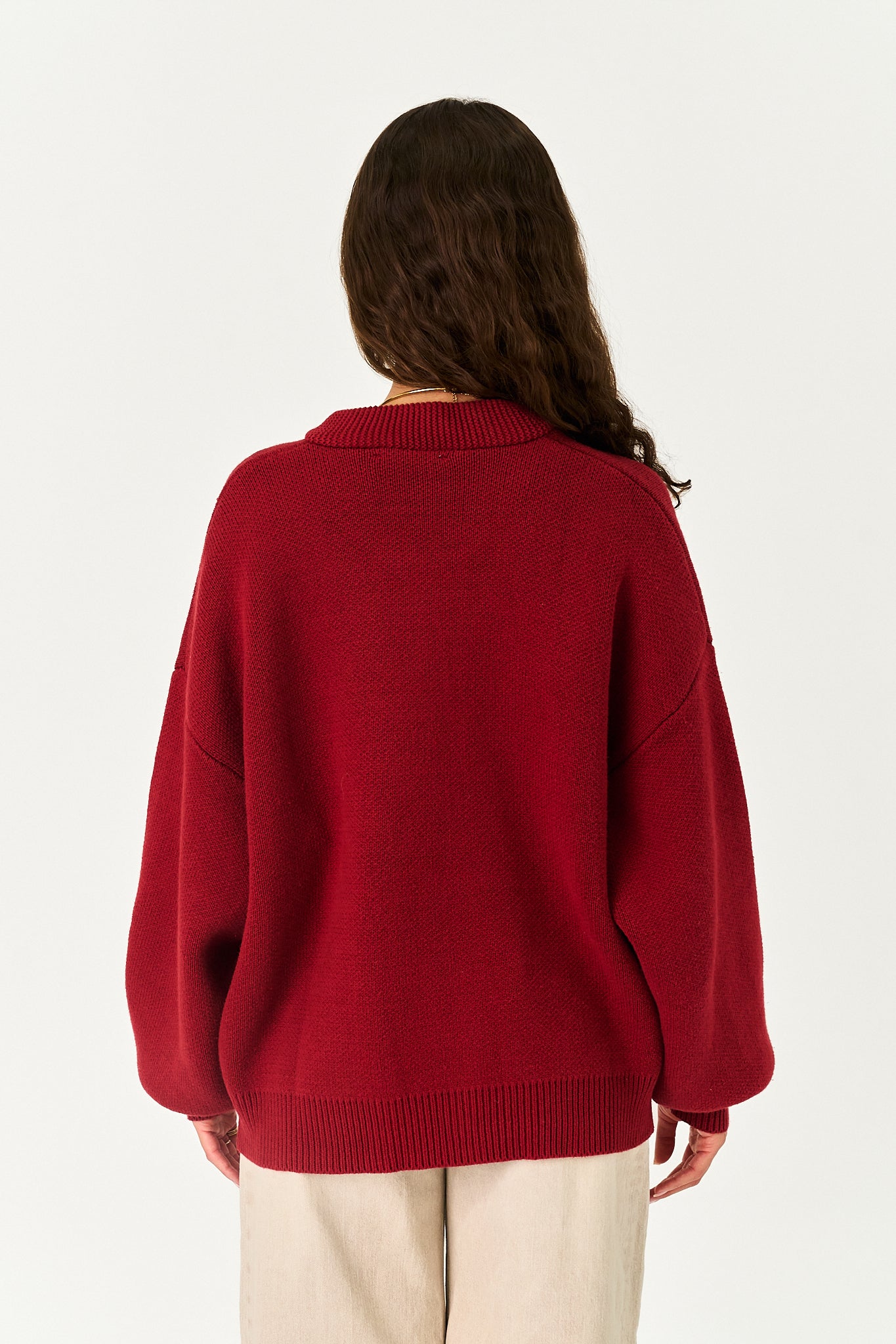 A woman viewed from behind wearing a deep red knit sweater and light beige pants against a plain white studio background.