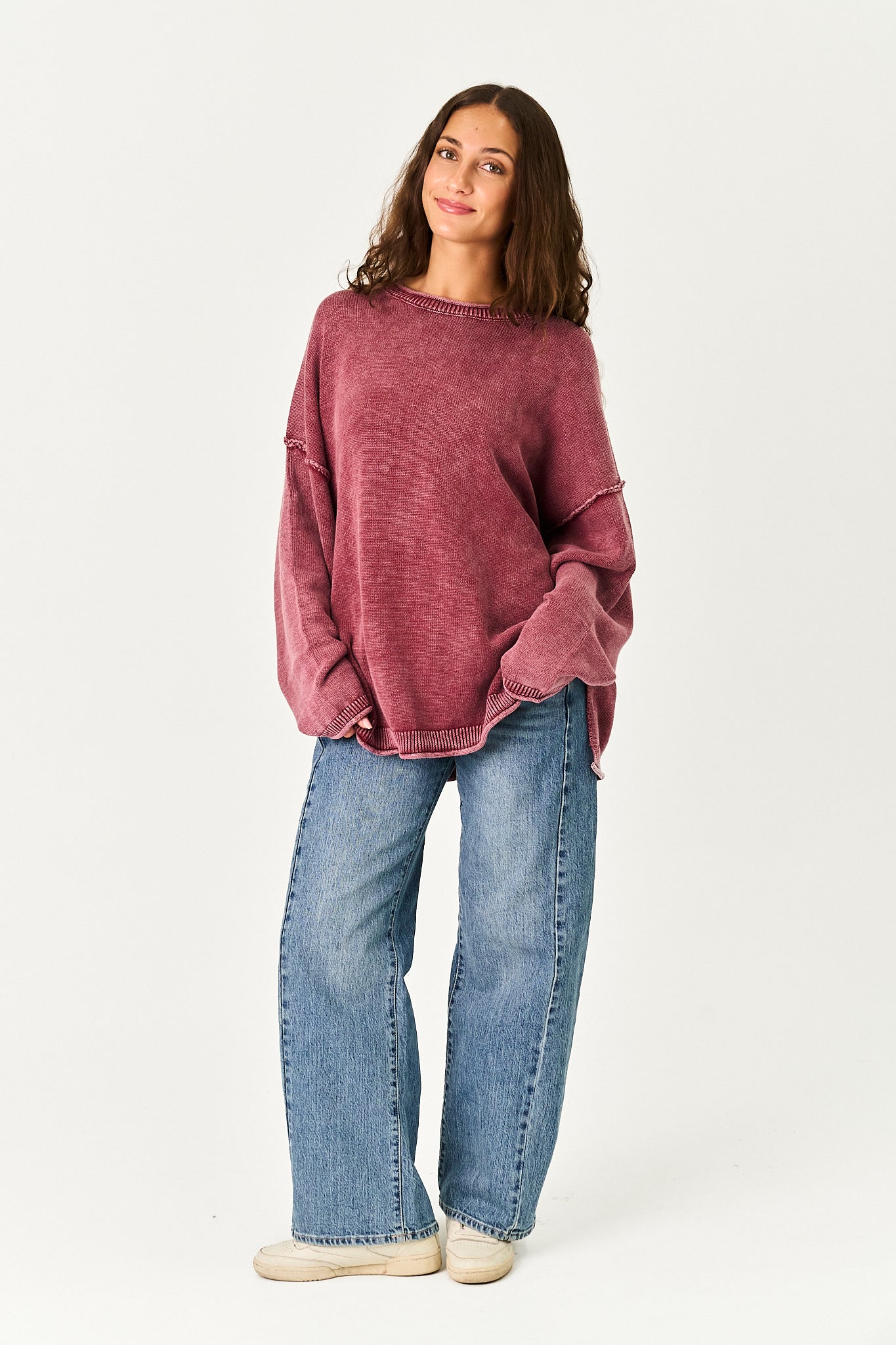 A woman modeling an oversized, washed-rose pink knit sweater paired with wide-leg blue jeans and beige sneakers, standing against a plain white studio background.