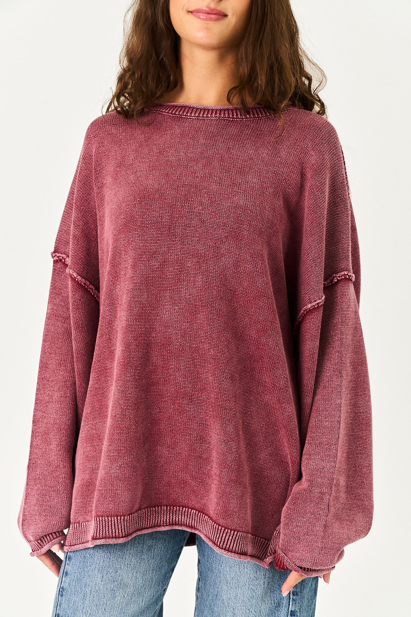 A woman wearing a washed, oversized knit sweater in a dusty rose or mauve color, paired with blue jeans, shown from the neck down against a plain light background.