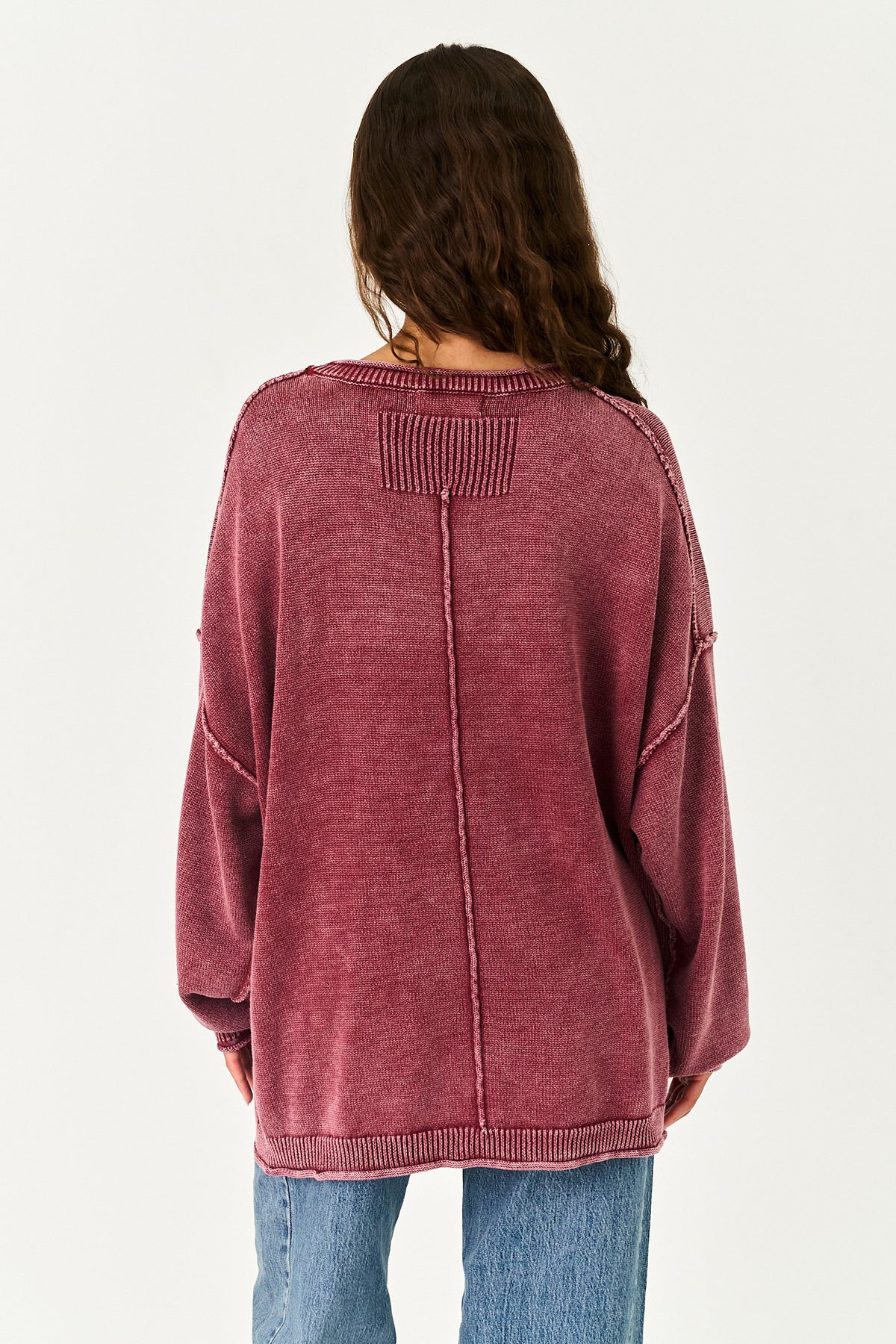 A woman viewed from the back wearing an oversized, washed-rose corduroy sweater and blue jeans against a plain white background.