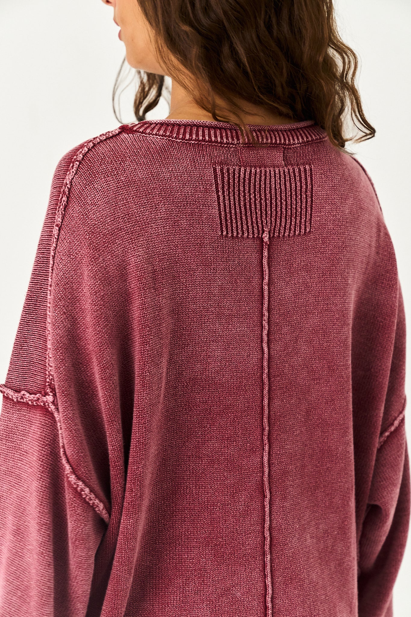 A close-up back view of a person wearing a textured, washed dusty rose knit sweater with unique seam details and a ribbed patch on the upper back.