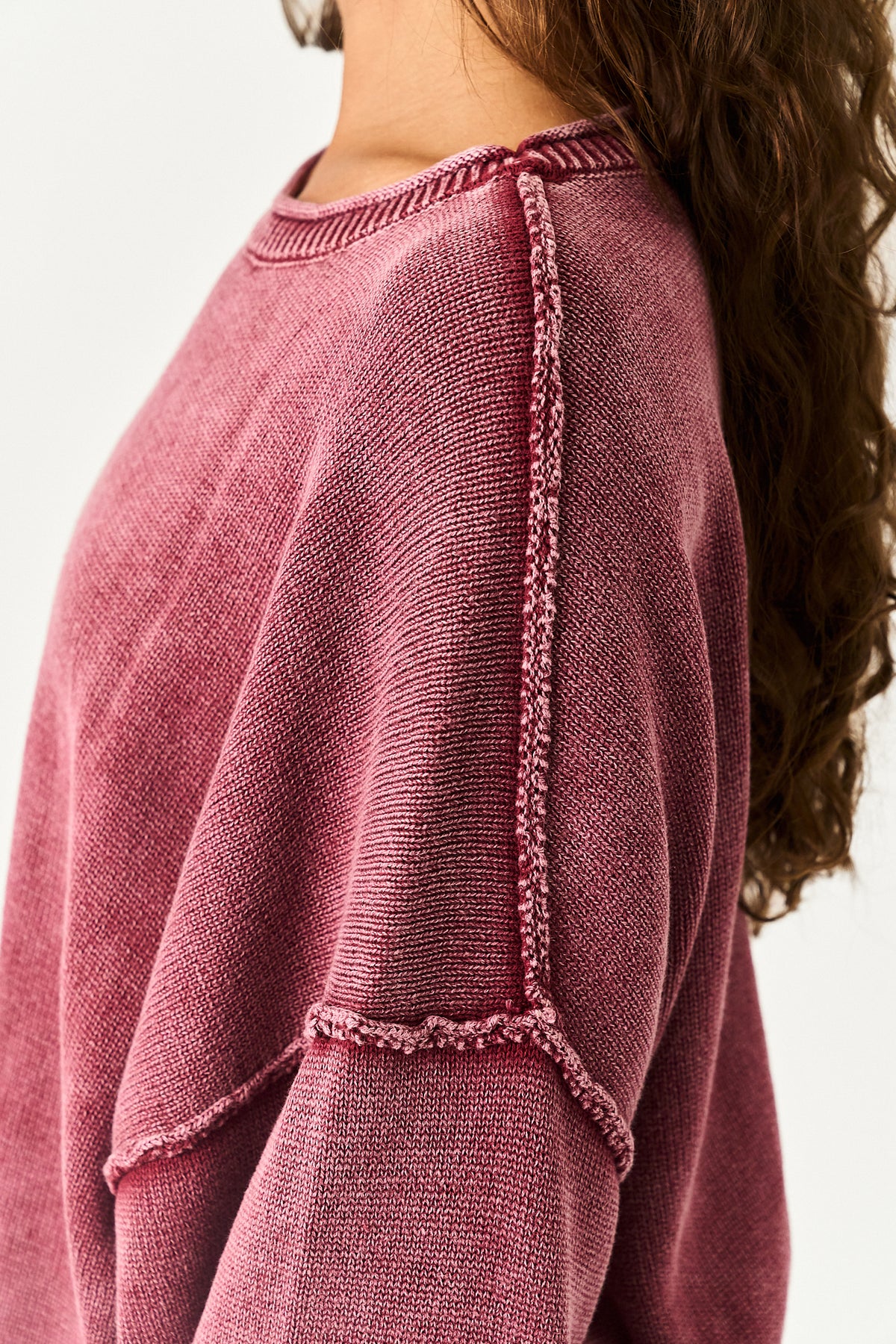 Close-up of a person wearing a textured knit sweater in a dusty rose or mauve pink color, showing the ribbed crew neck and decorative frayed seam details.