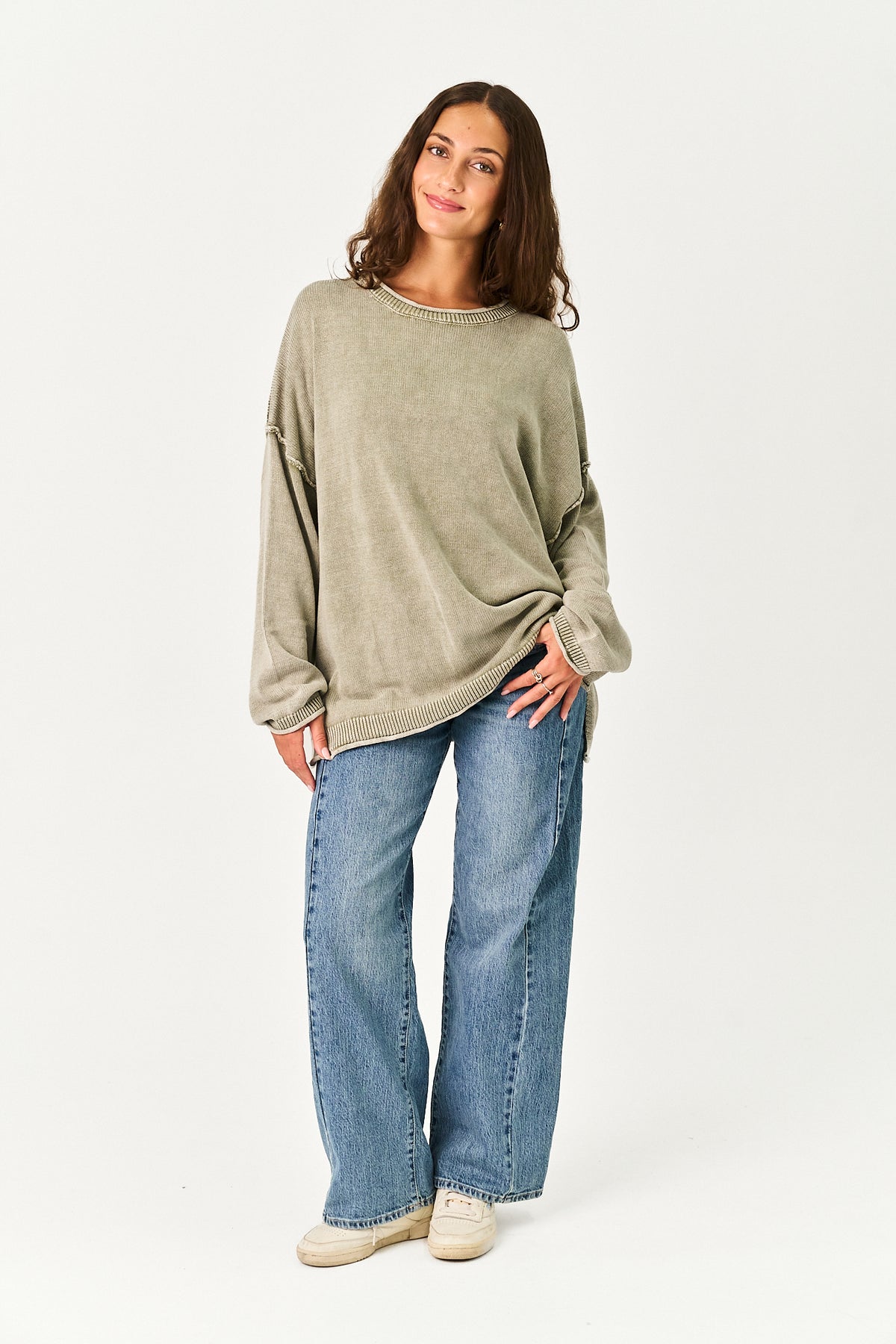 A woman modeling a loose, oversized sage green knit sweater paired with wide-leg blue jeans and white sneakers, standing against a plain white studio background.