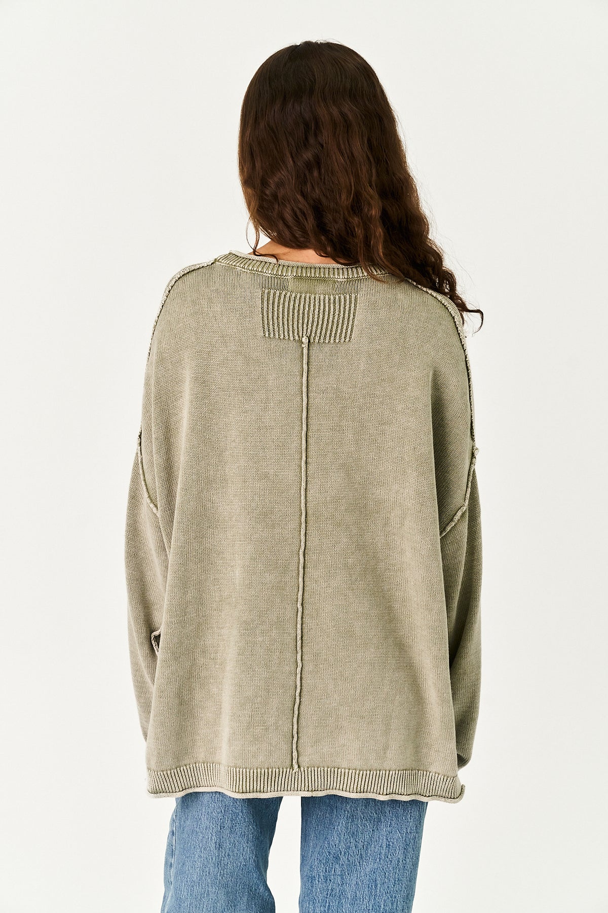 A woman viewed from behind wearing an oversized ribbed knit sweater in a muted sage green color, paired with blue jeans, against a plain light background.