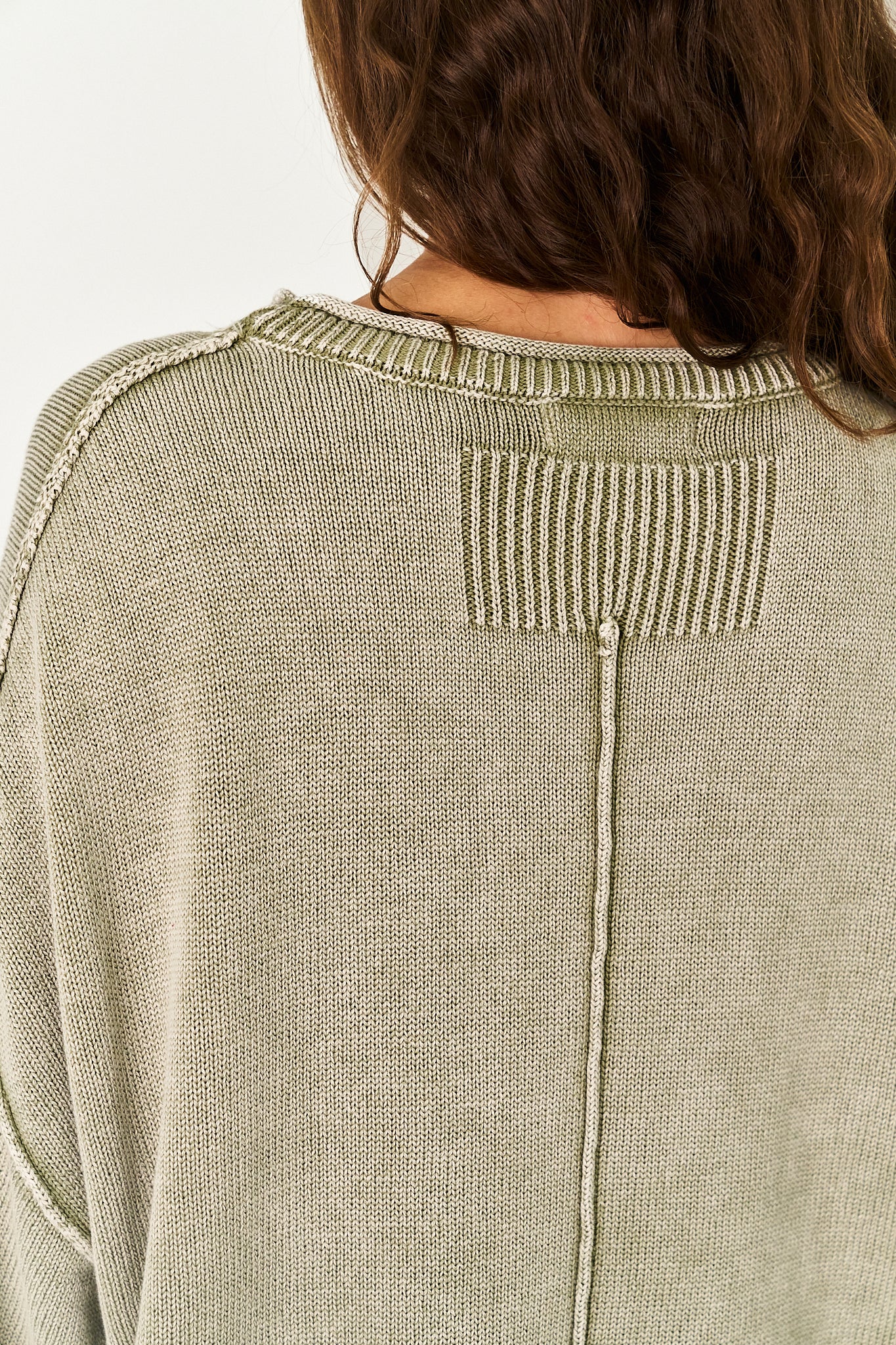 Close-up back view of a person wearing a light sage green knit sweater, showing the ribbed crew neck and a vertical seam detail.