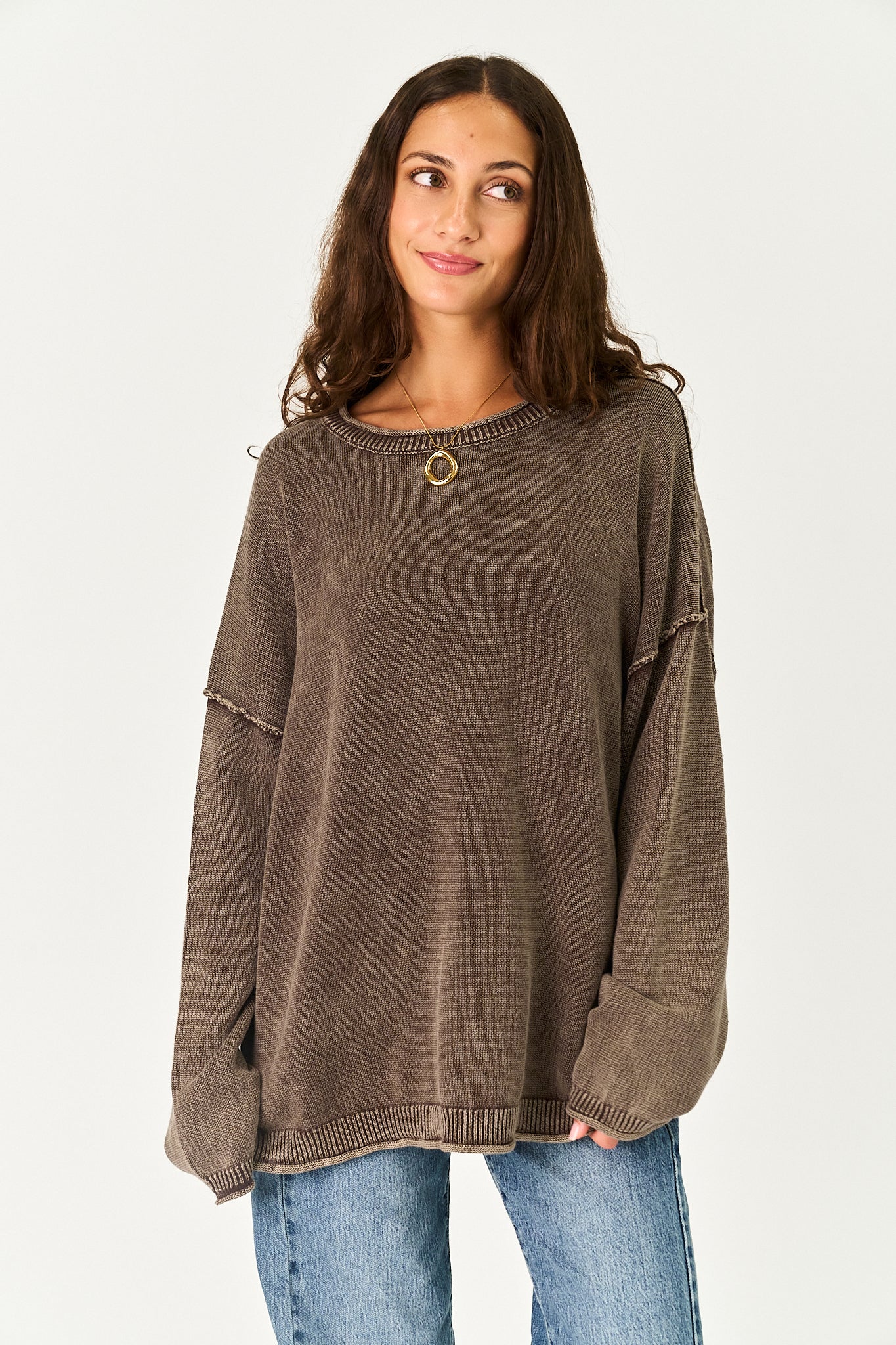 A woman wearing a brown textured knit sweater with a small gold ring detail at the neckline, paired with blue jeans, against a plain light background.