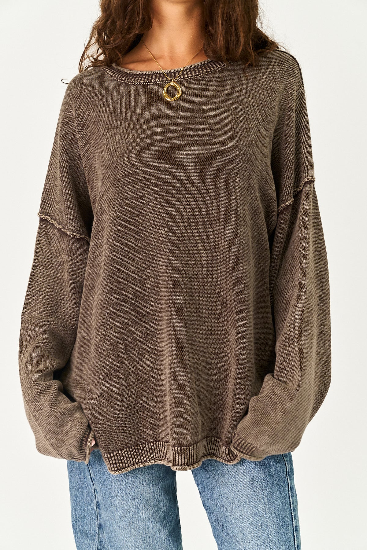A person wearing an oversized brown knit sweater with a small gold circular pendant at the neckline, paired with blue jeans, against a plain light background.