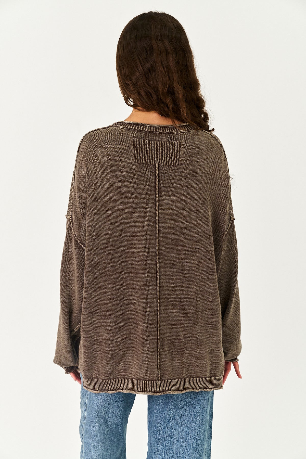 A woman viewed from the back wearing a brown ribbed knit sweater and blue jeans against a plain white background.
