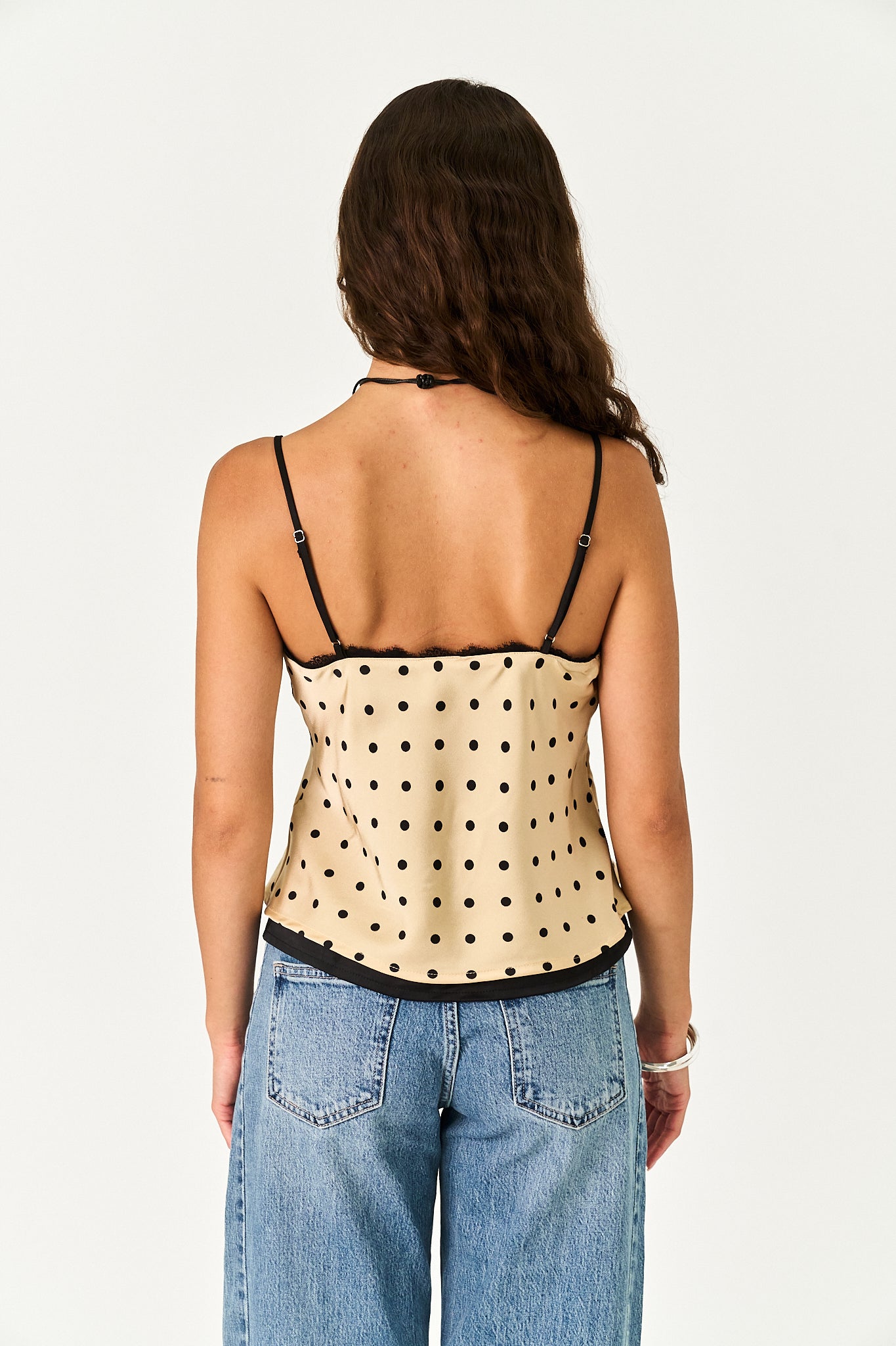 A woman shown from the back wearing a beige camisole top with black polka dots and thin black straps, paired with light blue jeans, against a plain white background.