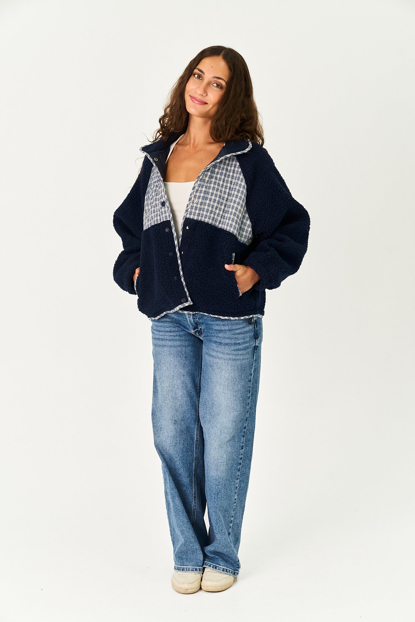 A woman modeling a navy blue fleece jacket with a gray and white gingham patterned yoke, paired with blue jeans and beige shoes, against a plain white studio background.