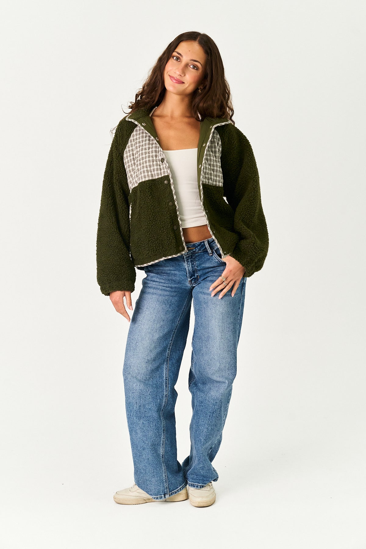 A woman modeling a dark green fleece zip-up jacket with beige and white gingham patterned panels on the chest, paired with blue jeans and white sneakers, against a plain white studio background.