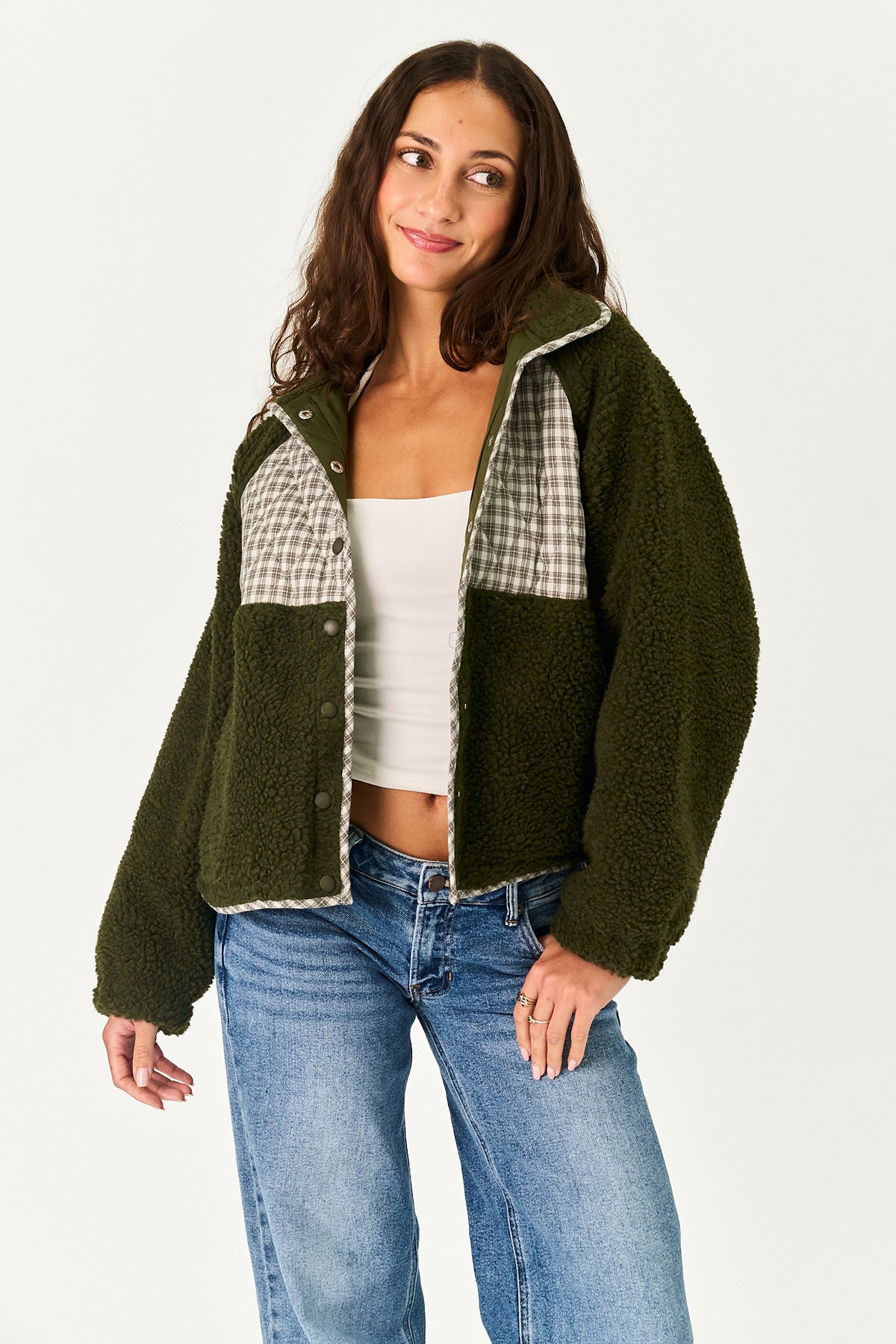 A woman modeling an olive green sherpa fleece jacket with beige and white gingham plaid panels on the chest, styled with a white tank top and blue jeans against a plain white studio background.