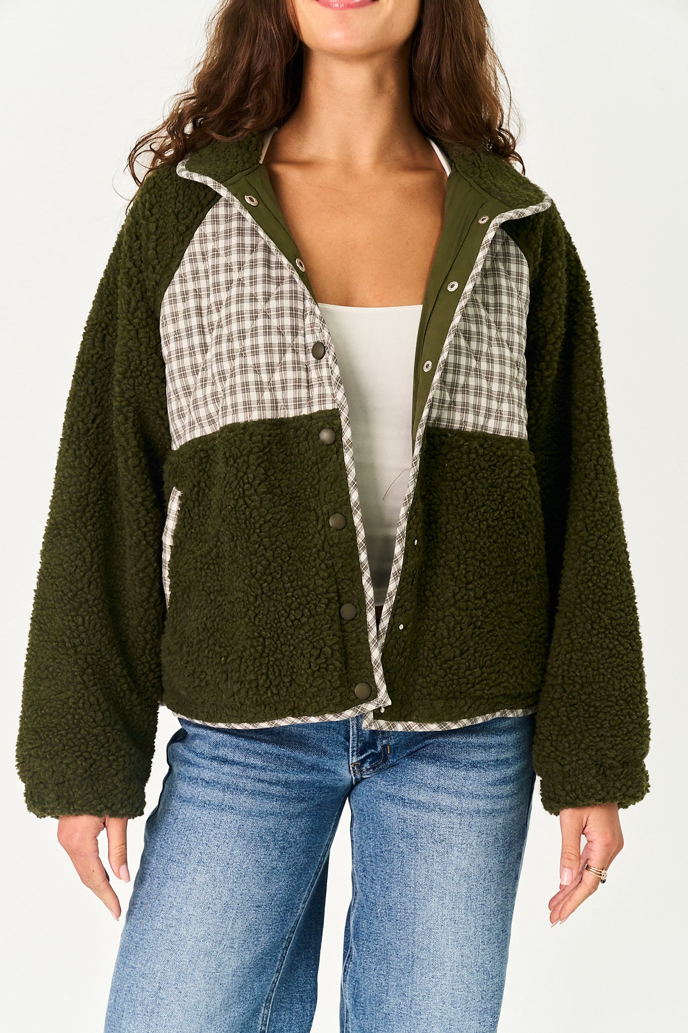 A woman wearing an olive green sherpa fleece jacket with beige and white gingham plaid panels on the chest, styled with a white tank top and blue jeans against a plain white background.
