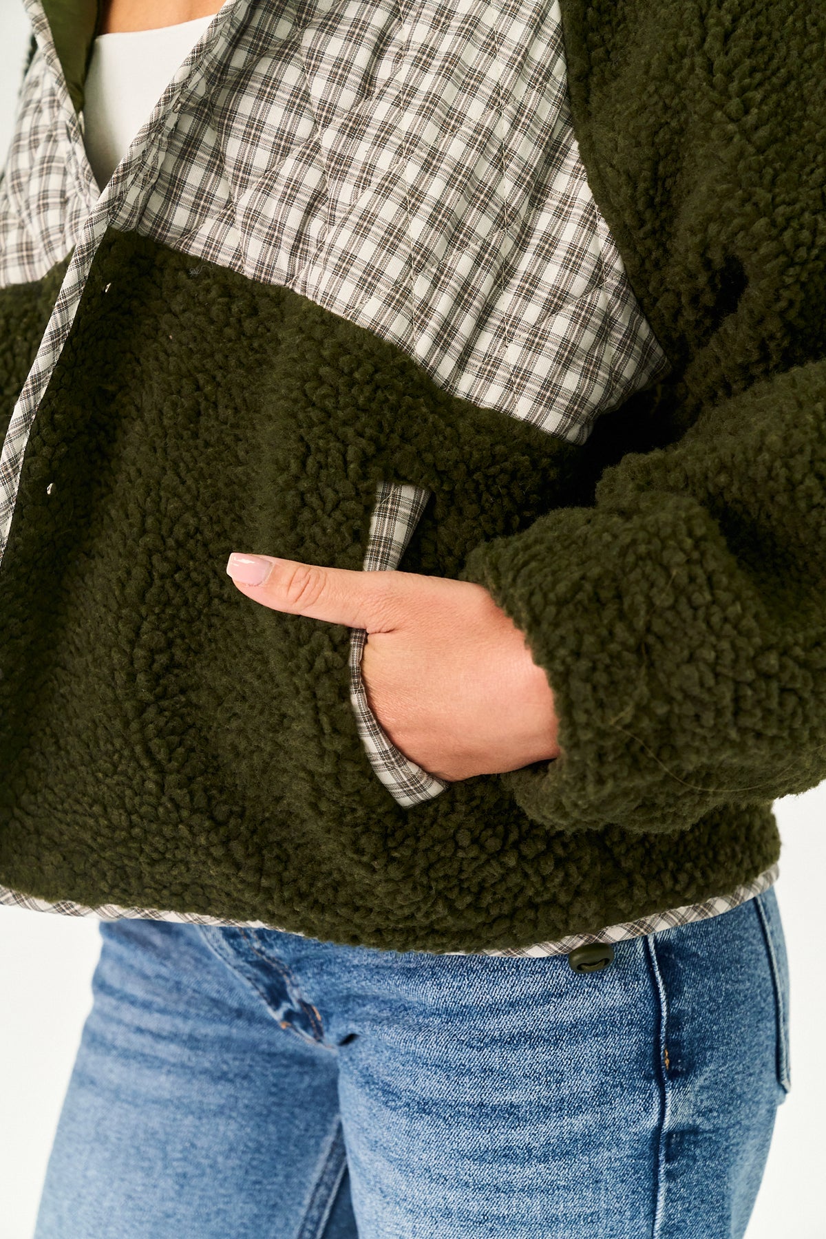 Close-up of a person wearing a color-block fleece jacket with an olive green sherpa body, a black and white plaid collar and shoulder panel, and blue jeans.
