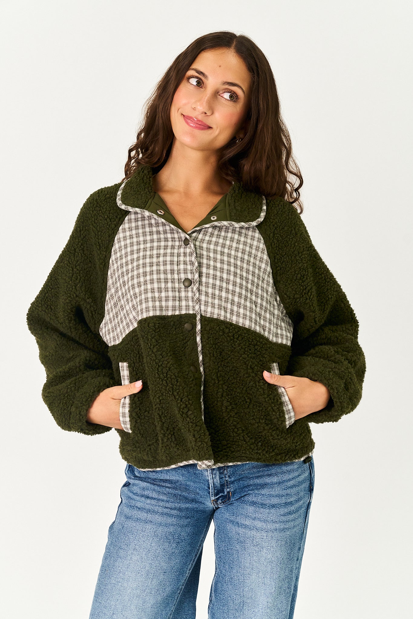 A woman wearing a dark olive green sherpa fleece jacket with a beige and white gingham check panel on the chest, paired with blue jeans, against a plain white studio background.
