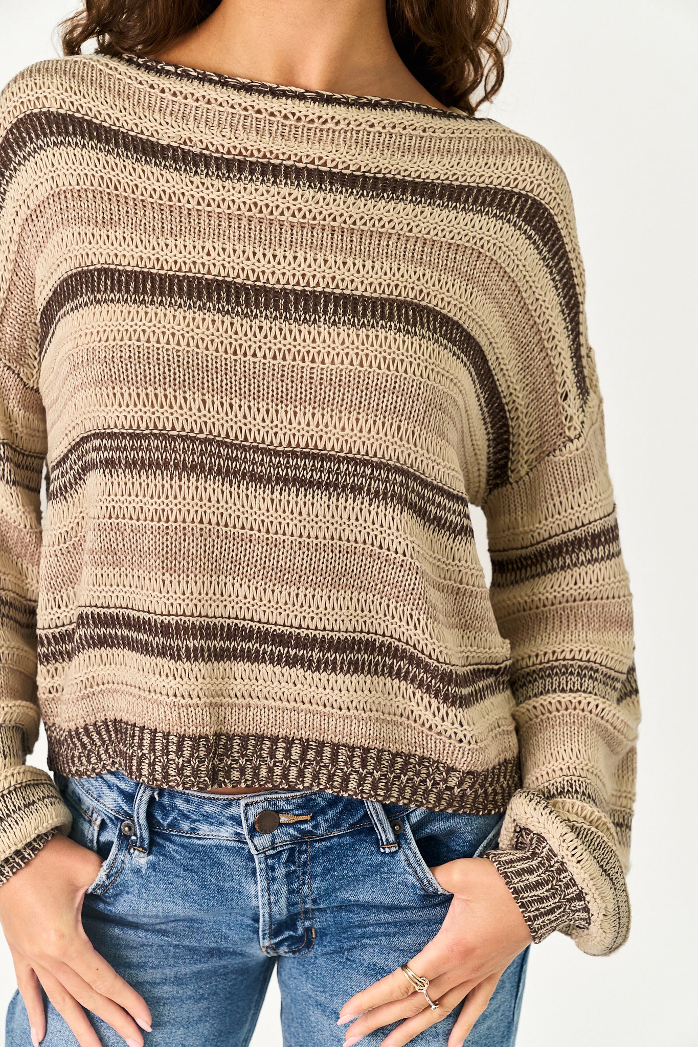 A woman wearing a beige and brown striped knit sweater with blue jeans, shown from the shoulders to the hips against a plain light background.