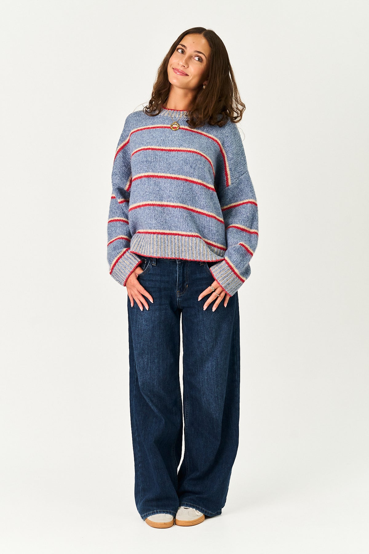 A woman modeling a blue knit sweater with red and white horizontal stripes, paired with dark blue wide-leg jeans, against a plain white studio background.