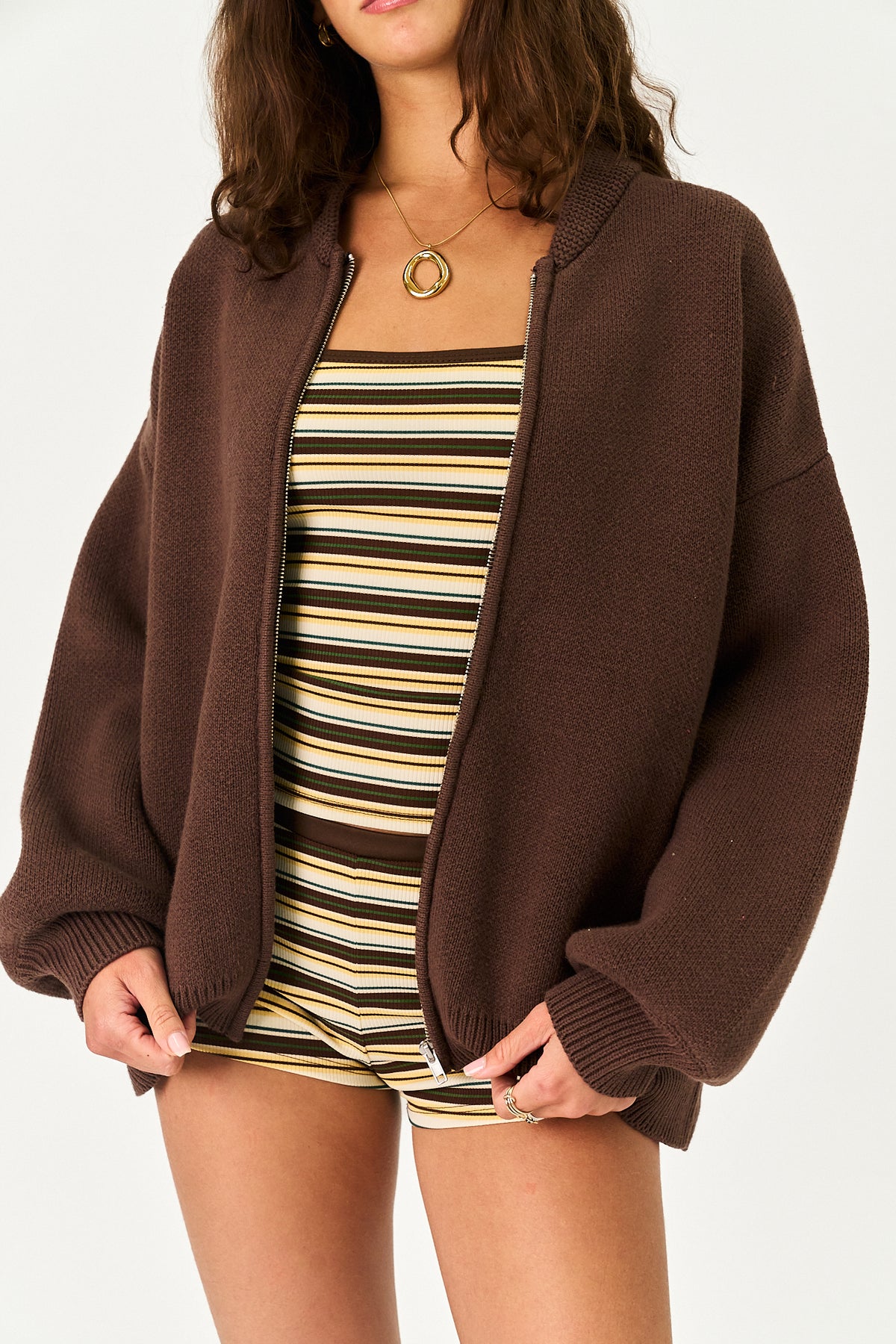 A woman wearing an oversized brown knit zip-up cardigan over a beige and brown striped romper, photographed against a clean white studio background.