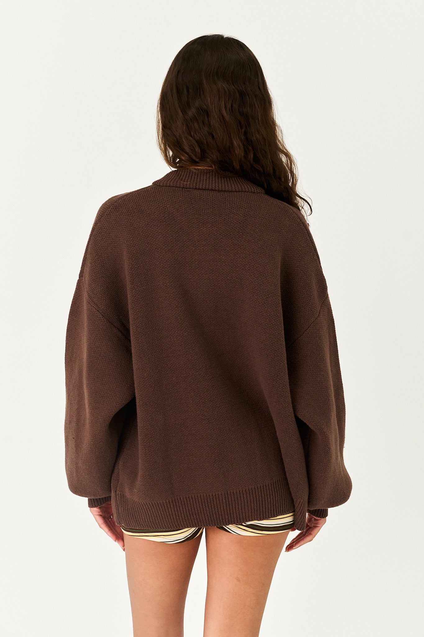 A woman viewed from behind wearing an oversized brown knit sweater and striped shorts against a plain light background.