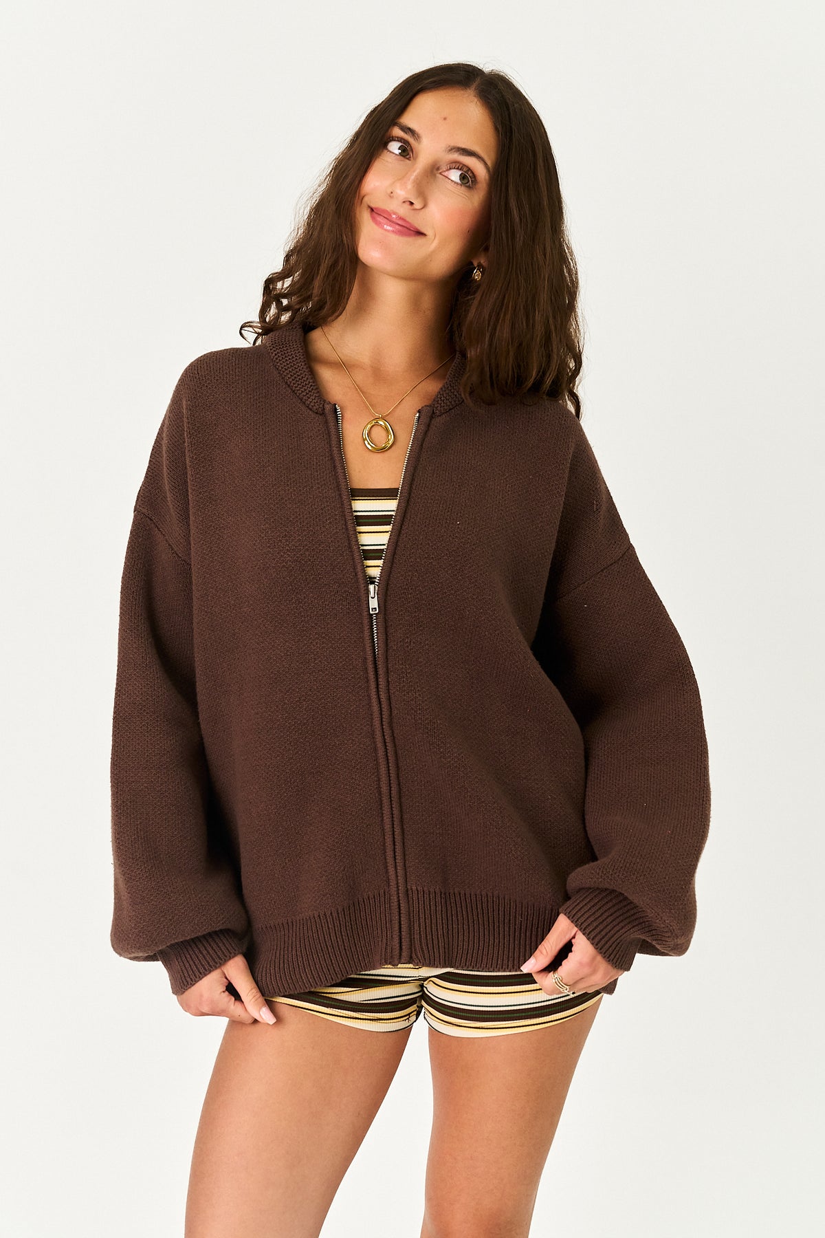 A woman wearing an oversized brown zip-up knit sweater over a striped top and striped shorts, photographed against a plain light gray studio background.