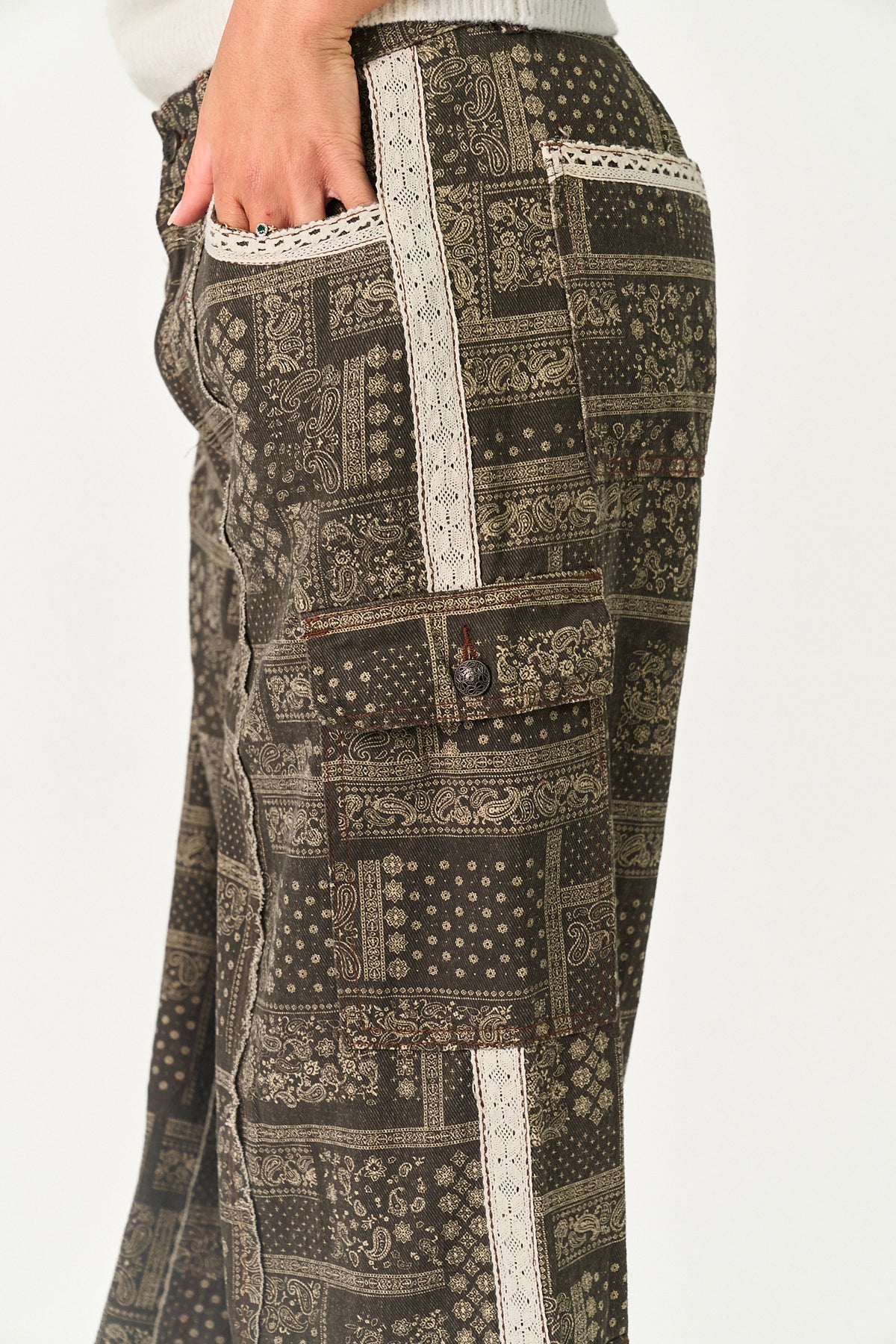 Close-up of a person wearing patterned cargo pants with a dark brown and beige patchwork print and white lace trim, shown against a plain light background.