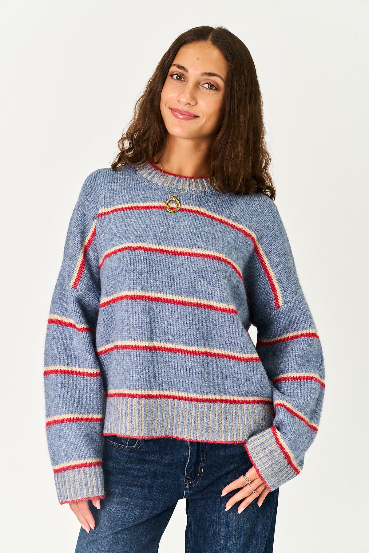 A woman wearing a blue knit sweater with horizontal red and cream stripes, paired with dark blue jeans, against a plain light gray studio background.