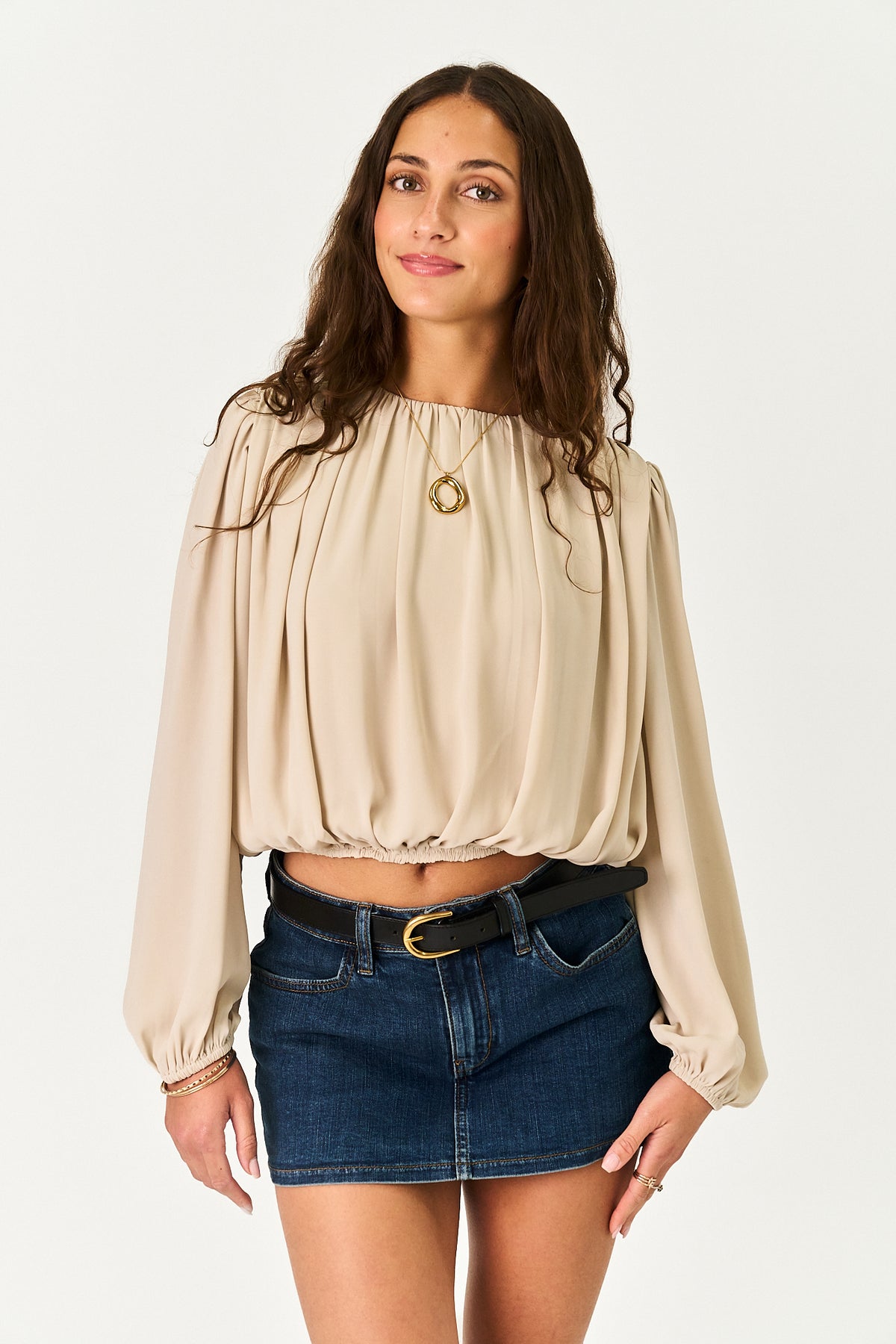 A woman modeling a beige long-sleeve blouse with a gathered, bubble-style neckline and cuffs, paired with a dark blue denim mini skirt and a black belt, against a plain white studio background.