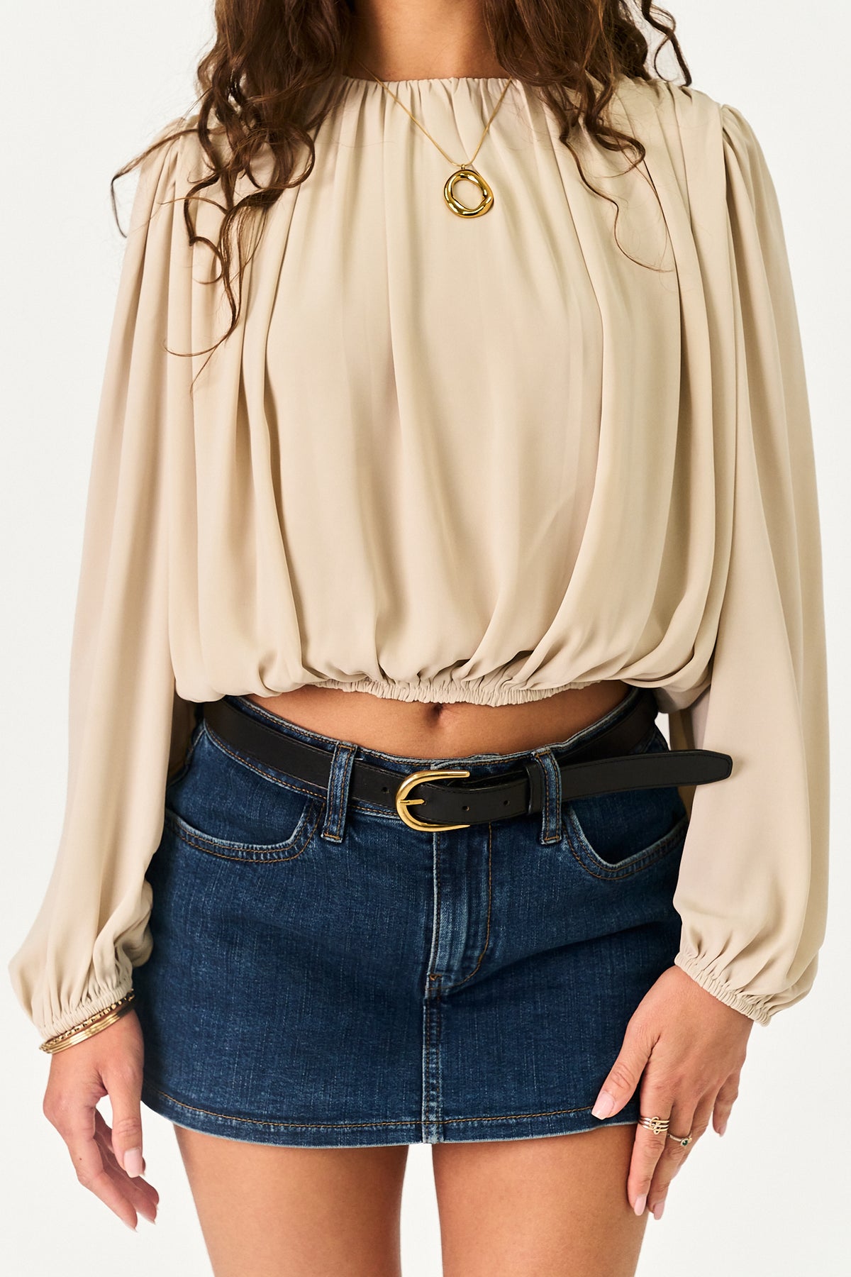 A woman wearing a beige long-sleeve blouse with a gathered, balloon-like silhouette and a high neckline, paired with a dark blue denim mini skirt and a black belt with a gold buckle.